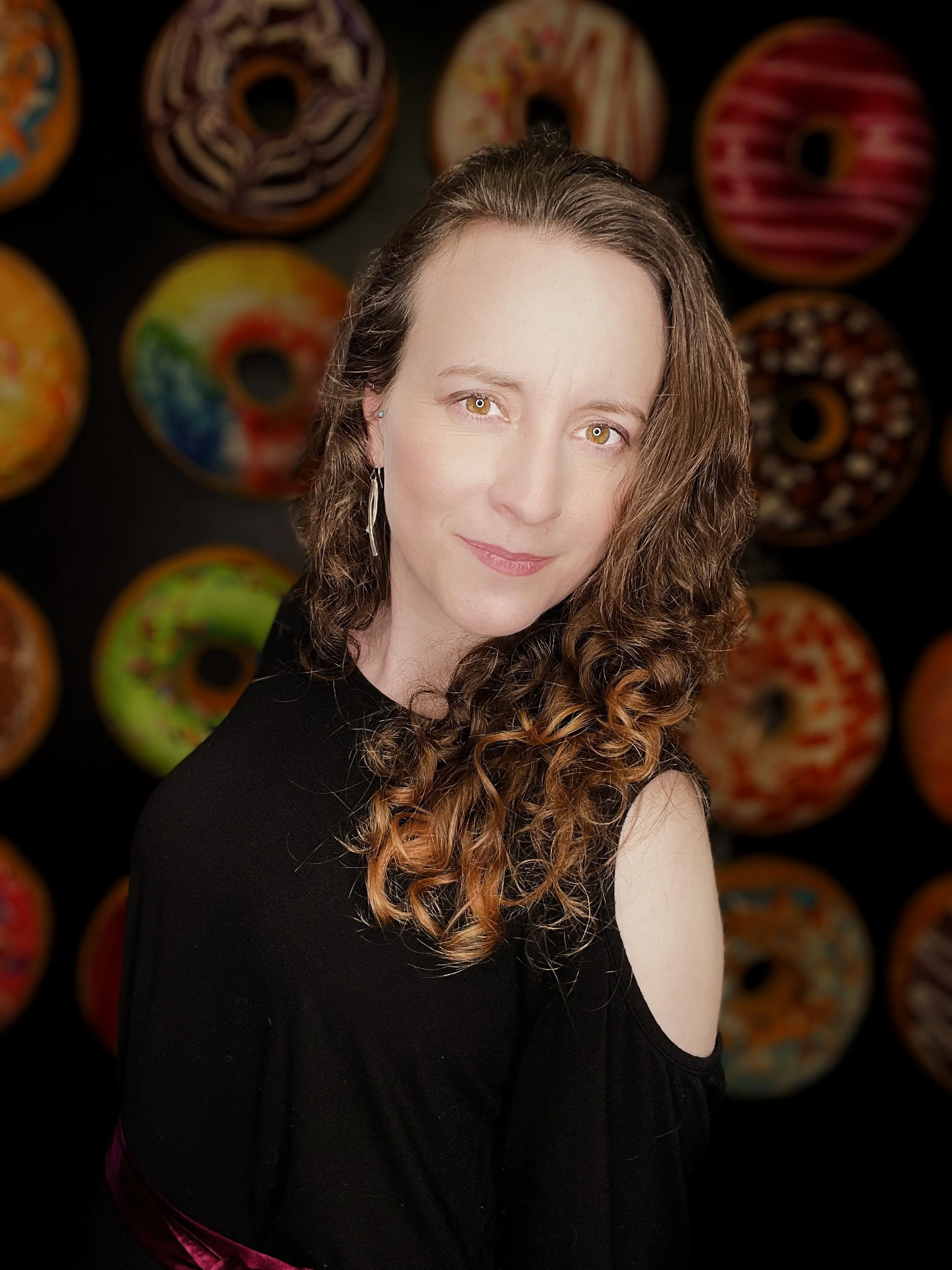 A woman with long curly brown hair and light skin, wearing a black top with cutout shoulder, standing in front of a background of colorful, decorated donuts.