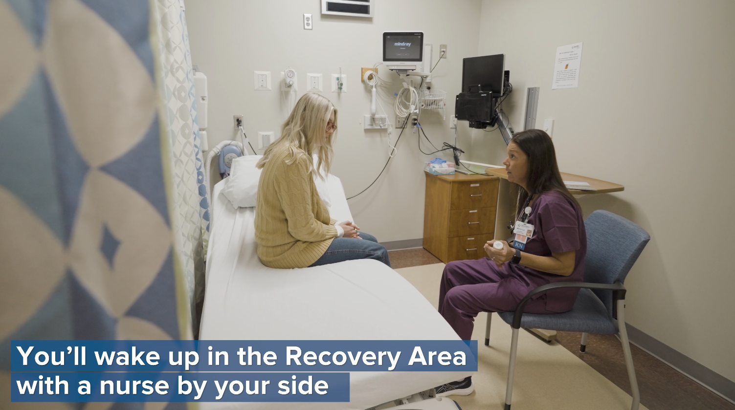 A nurse speaking with a seated person in a clinical room.
