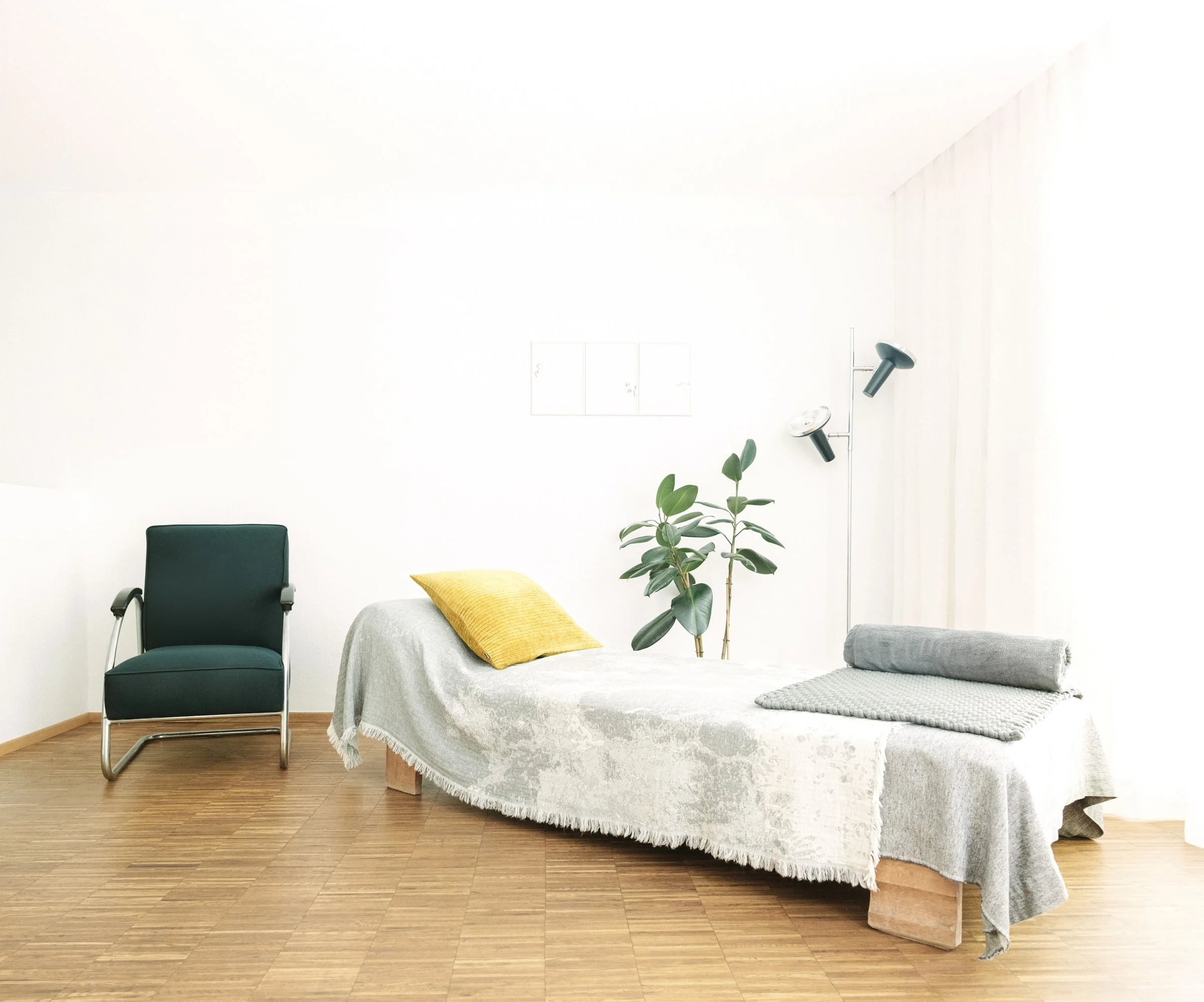 Ein schlicht eingerichteter Raum mit einem Bett, einem schwarz-gelben Sessel, einer Zimmerpflanze und einem Leselicht mit drei Lampen, alles auf einem Holzboden.