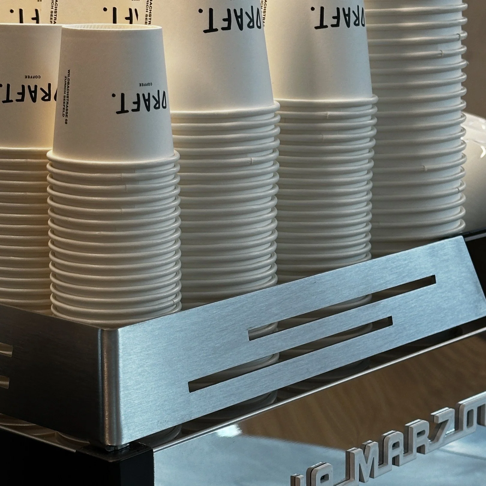 A stack of white paper cups with the word 'DARK' printed on them, placed inside a metallic container with slots, next to a stack of white plates.