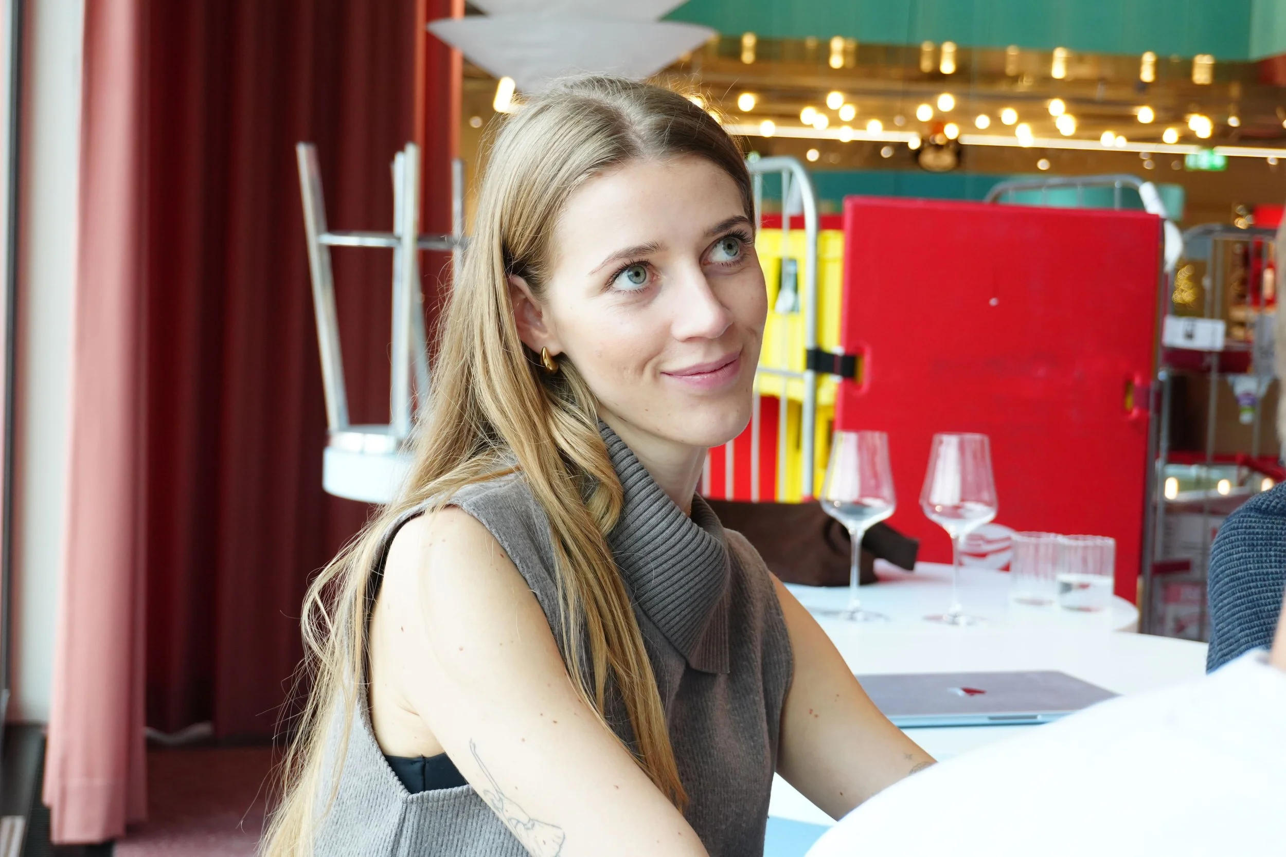 A woman with long blonde hair and blue eyes sitting at a table in a cafe or restaurant, smiling and looking upward. There are glasses and a menu on the table behind her, with blurred colorful lights and structures in the background.
