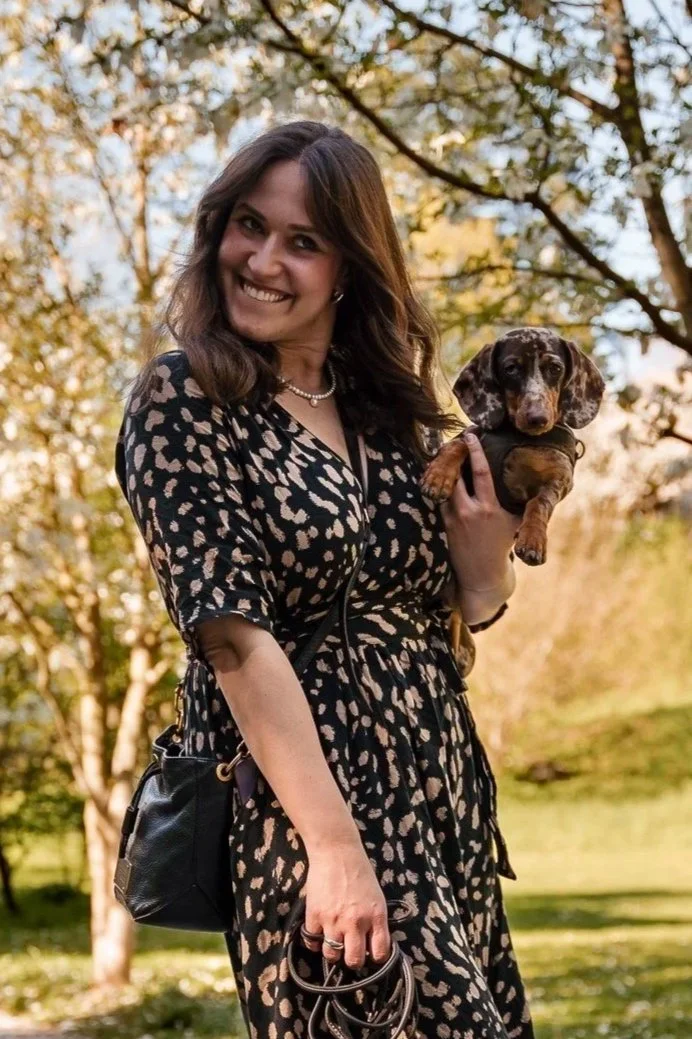 A woman in a patterned dress holding a puppy outdoors with trees in the background.