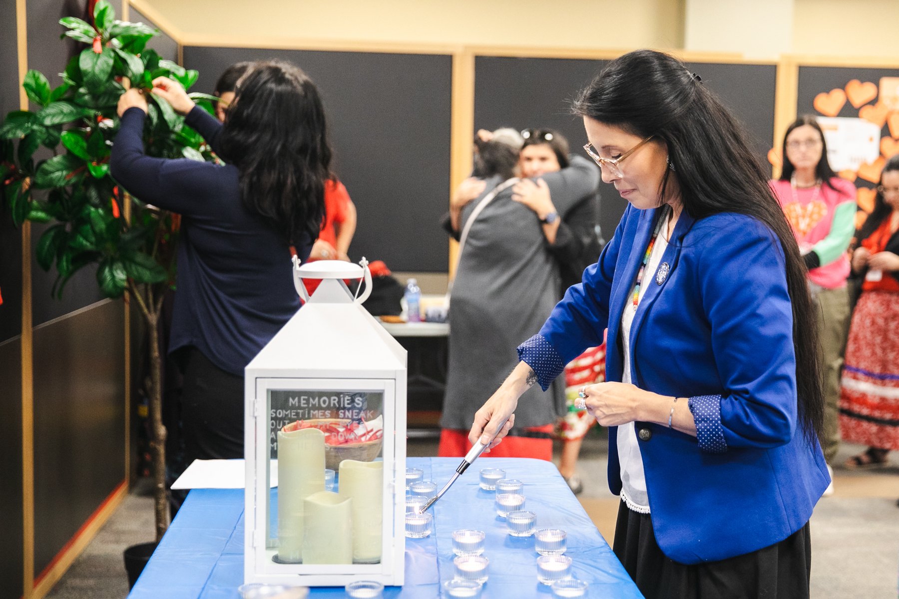 A woman in a blue blazer lights small candles on a table during a memorial or remembrance event. Other people in the background are hugging and socializing.