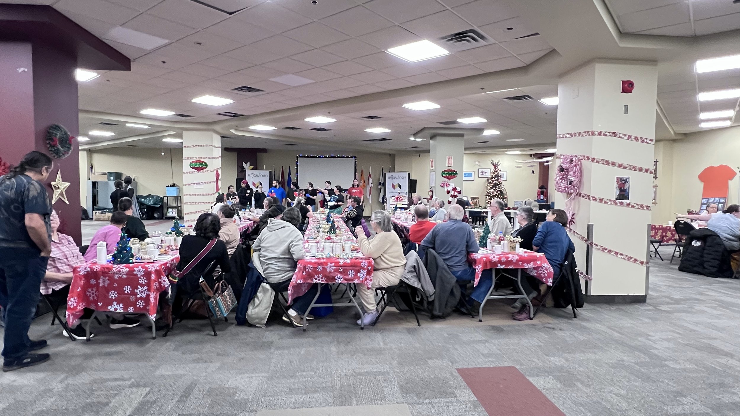 A large indoor Christmas celebration with people sitting at decorated tables with red snowflake tablecloths, Christmas trees, and holiday decorations, watching a stage with performers.