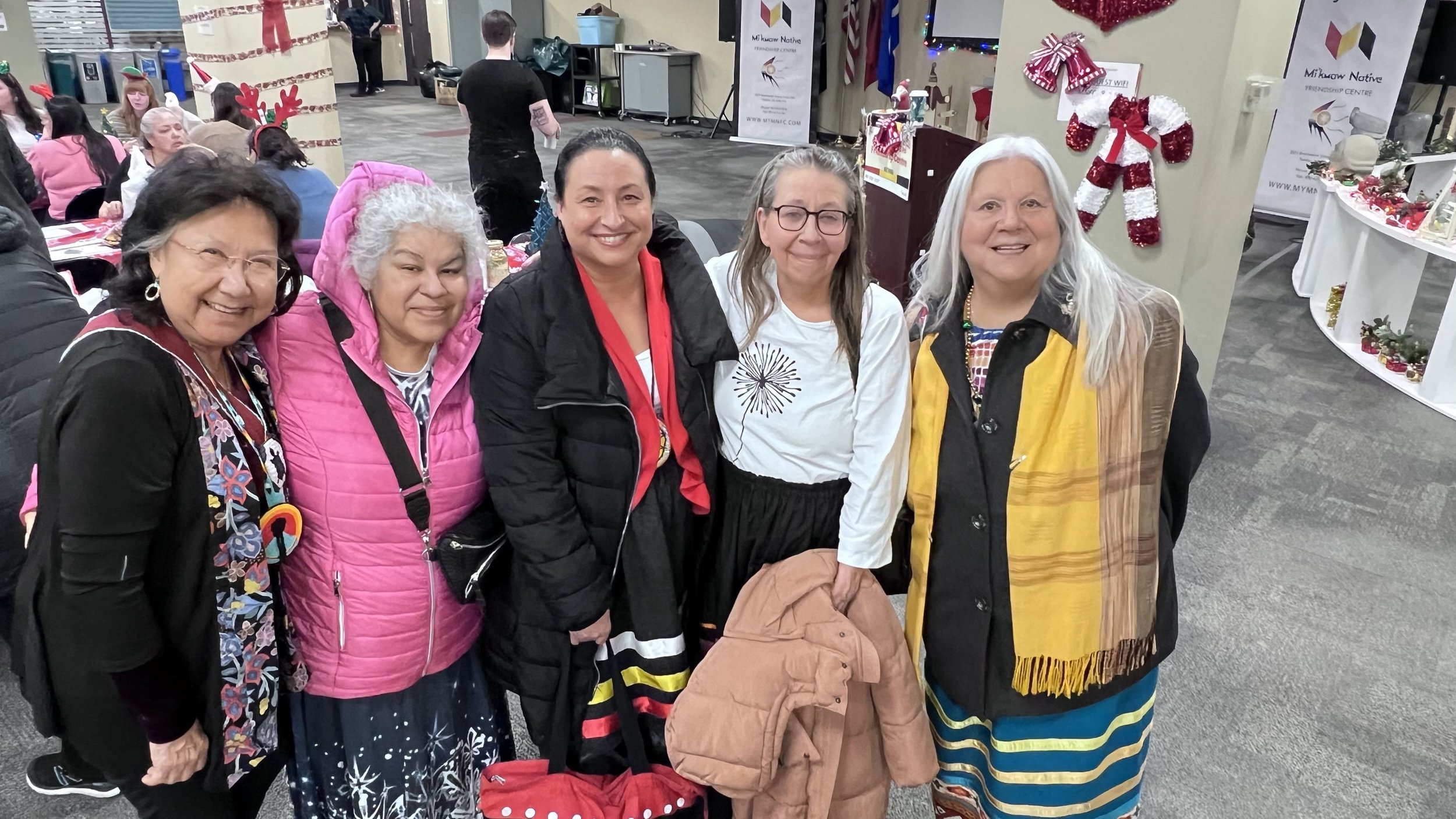 Five women standing together at a Christmas event. They are smiling, wearing colorful clothing and accessories. The background features decorated holiday ornaments, tables with Christmas displays, and other guests sitting and standing.