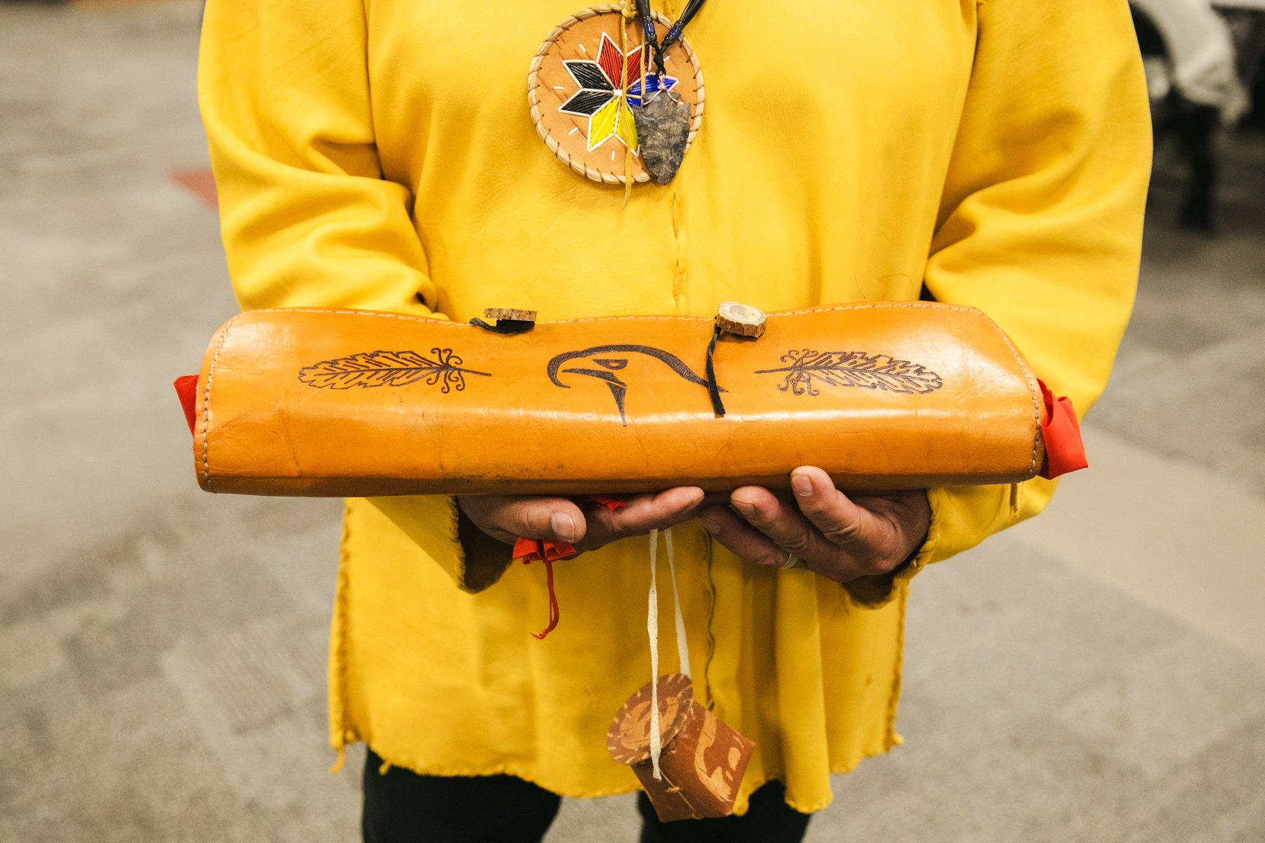 Person dressed in a yellow garment holding a wooden object decorated with black line drawings of leaves and a bird's head.