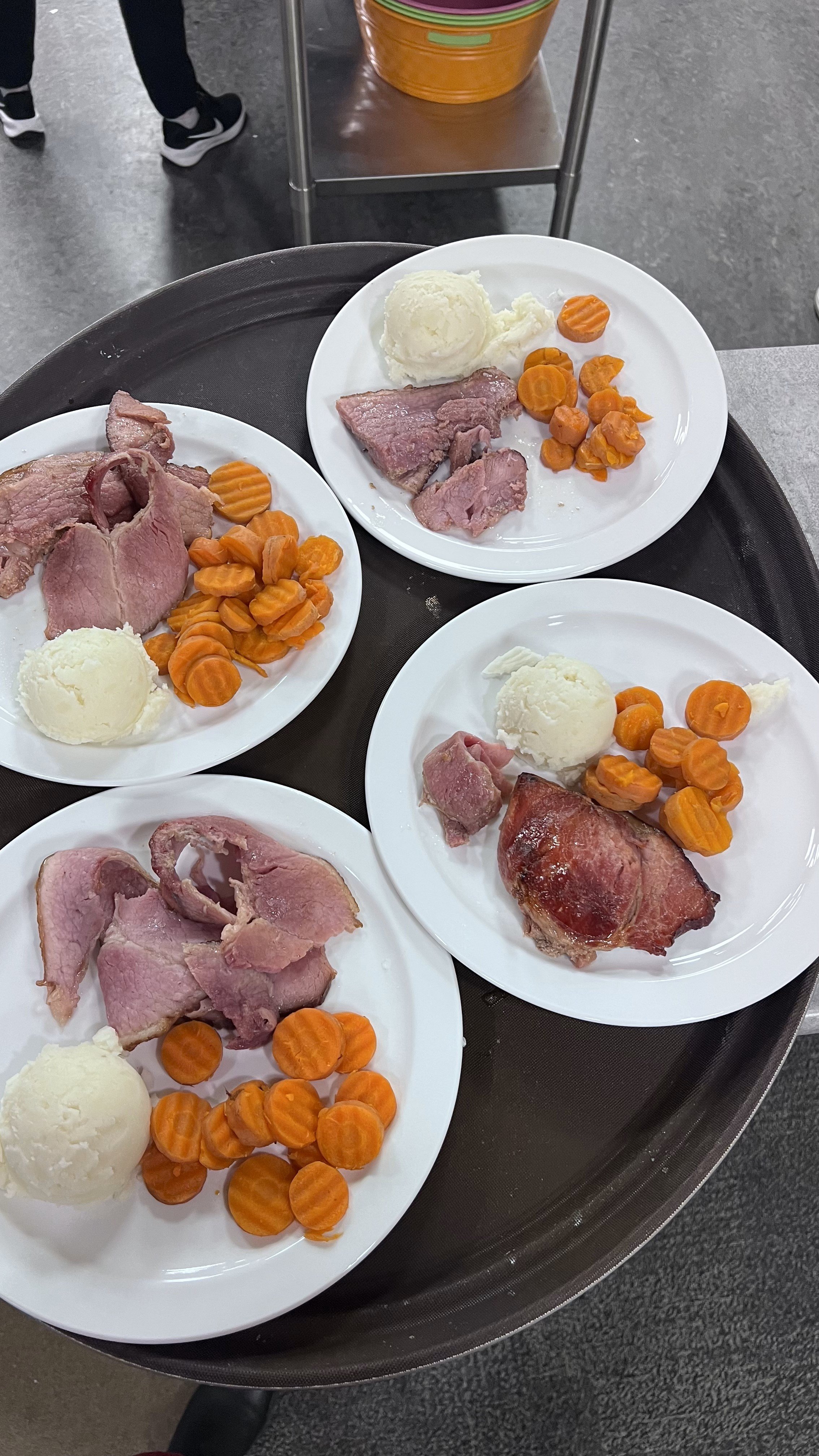 Four plates of cooked meats, mashed potatoes, and sliced carrots on a black tray, with a person’s legs and part of a cart in the background.