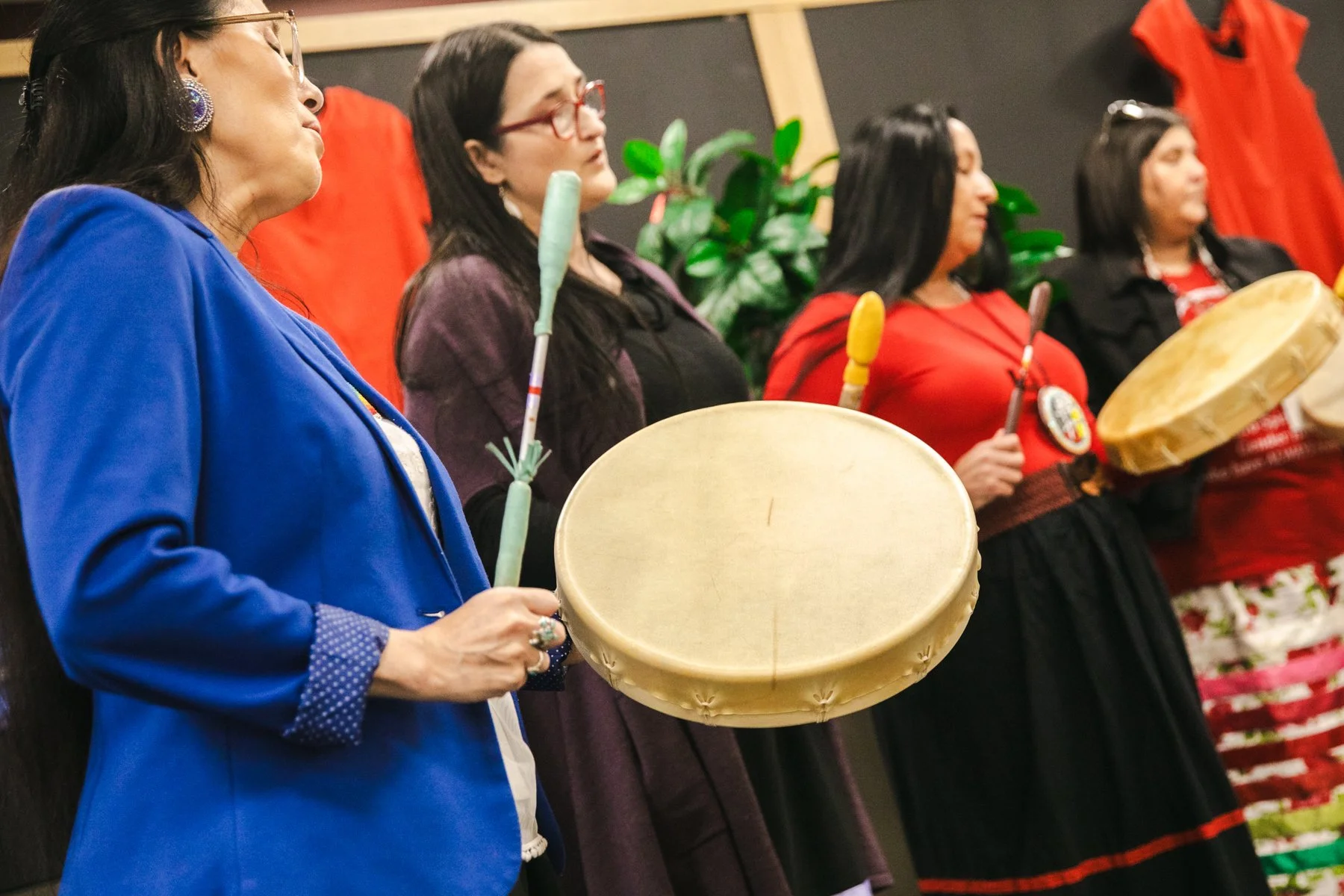 Four women standing in a row playing hand drums, with red and green decorations in the background.