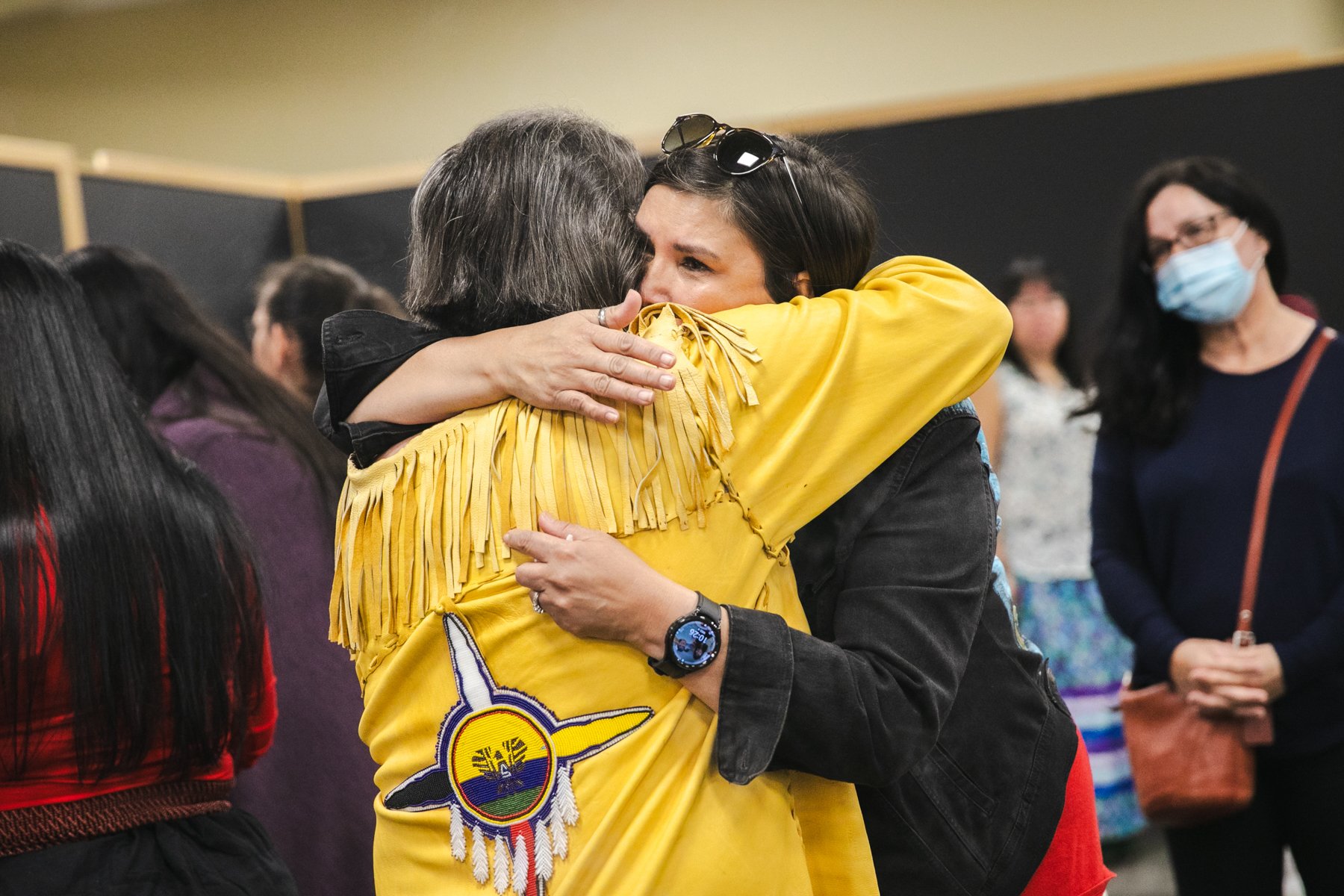 Two women hugging warmly in a crowded room, one wearing a yellow shirt with a colorful patch, and the other wearing a black jacket. In the background, other people are present, with one woman wearing a face mask.