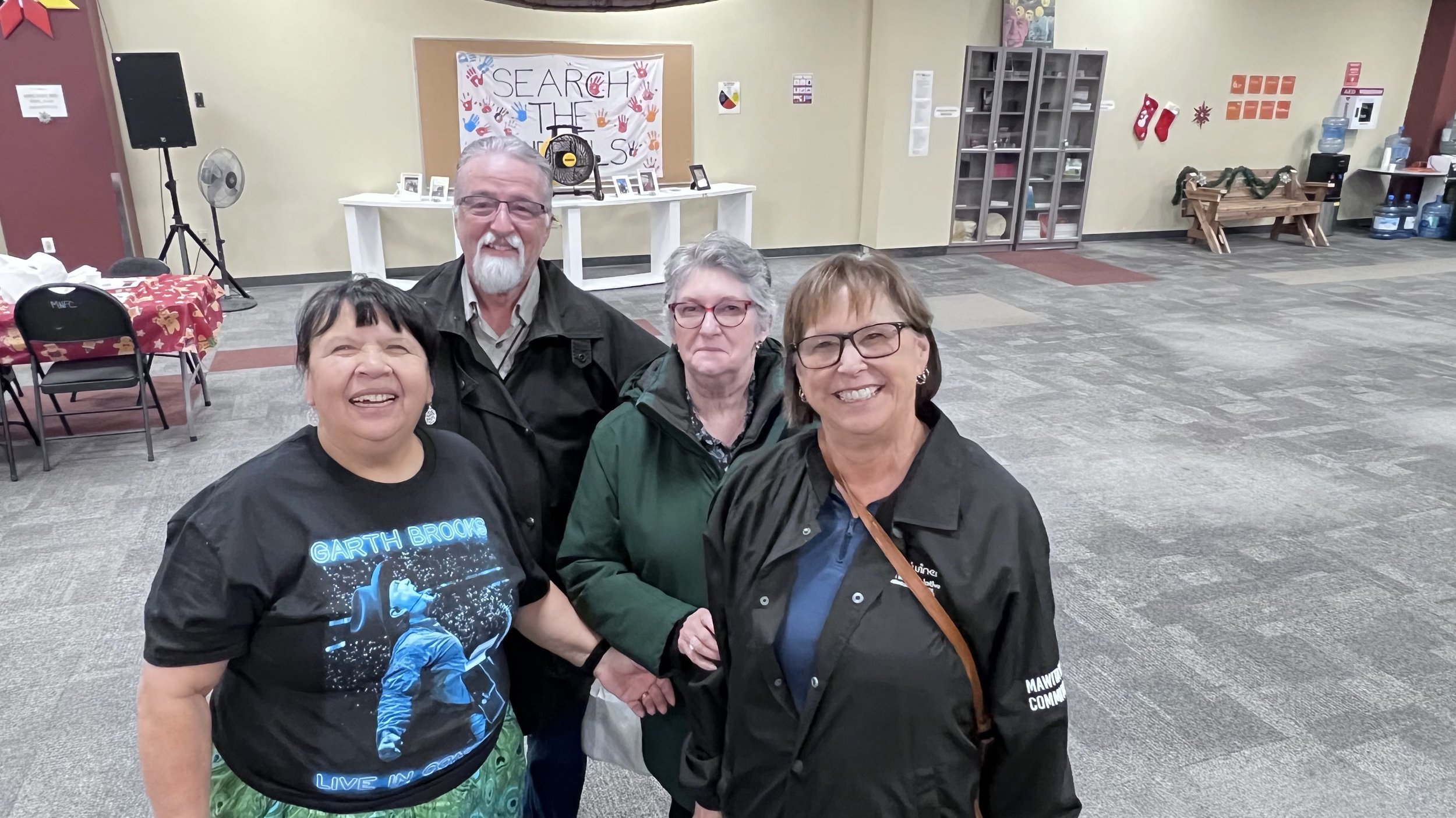 Four smiling adults standing together inside a community center decorated for the holidays, with a sign saying 'Search the Stars' and holiday decorations visible in the background.