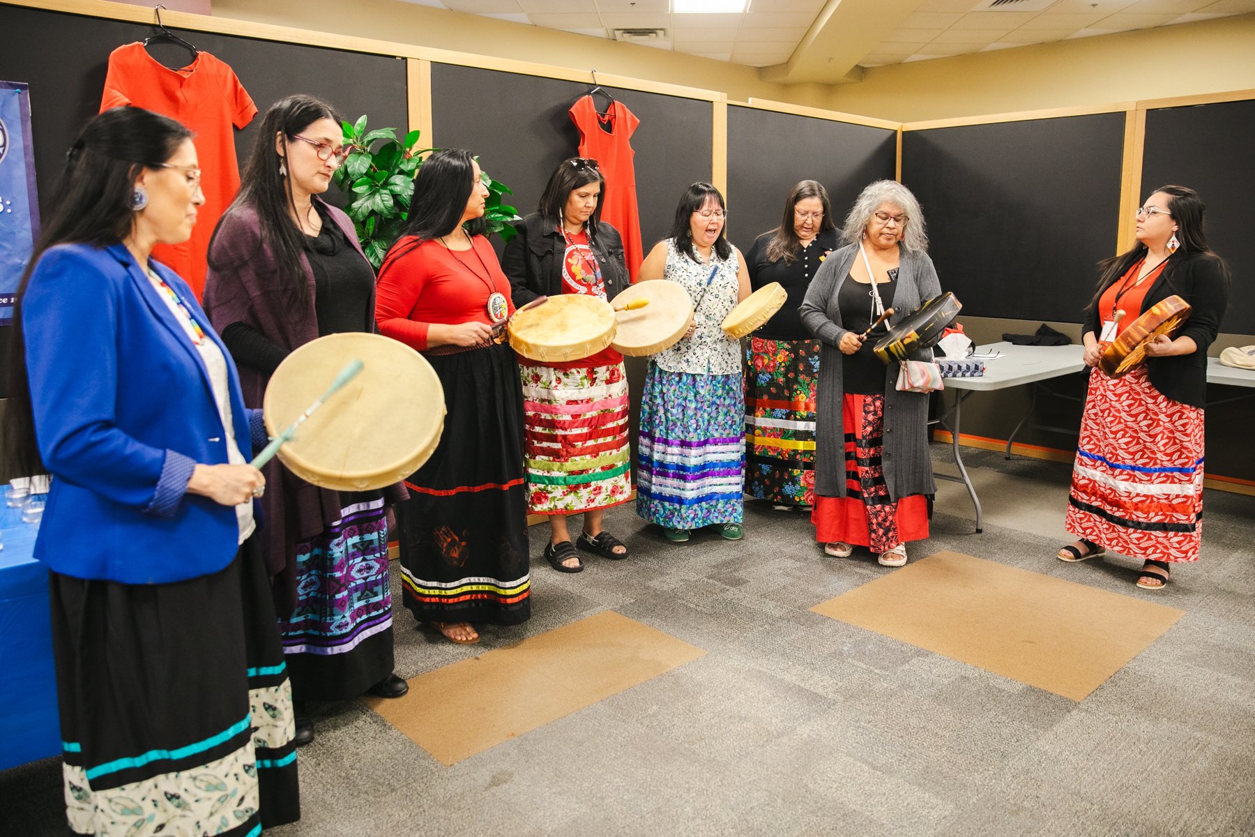 Group of women dressed in colorful traditional clothing playing hand drums in a circle at an indoor event.