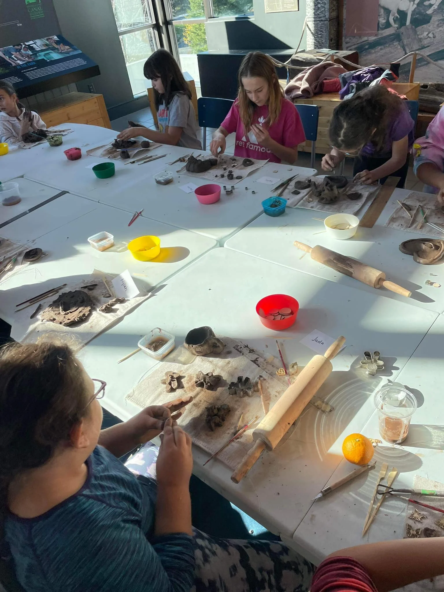 Children and adults engaging in pottery or clay sculpting activities around a white table with various clay pieces, tools, bowls, and rolling pins, in a well-lit room with large windows.