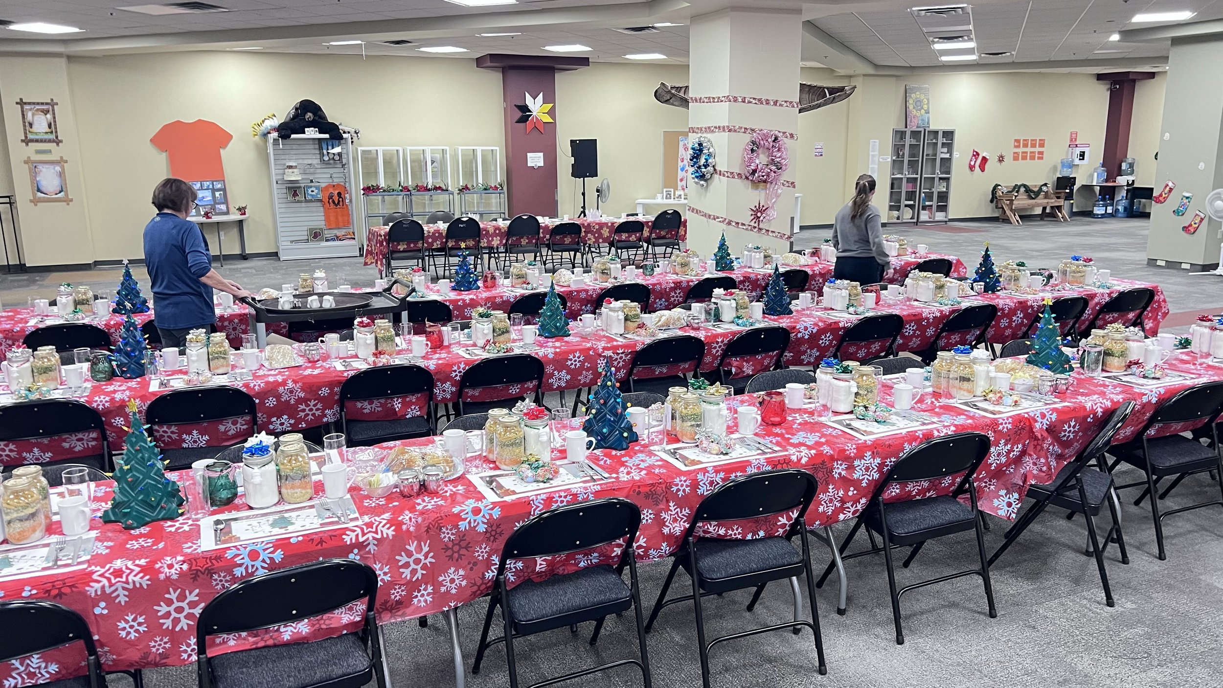 Decorated banquet hall with long tables covered in red snowflake tablecloths, set for a holiday event with small Christmas trees, jars, and cups. Two people are preparing the tables, with shelves and decorations visible in the background.