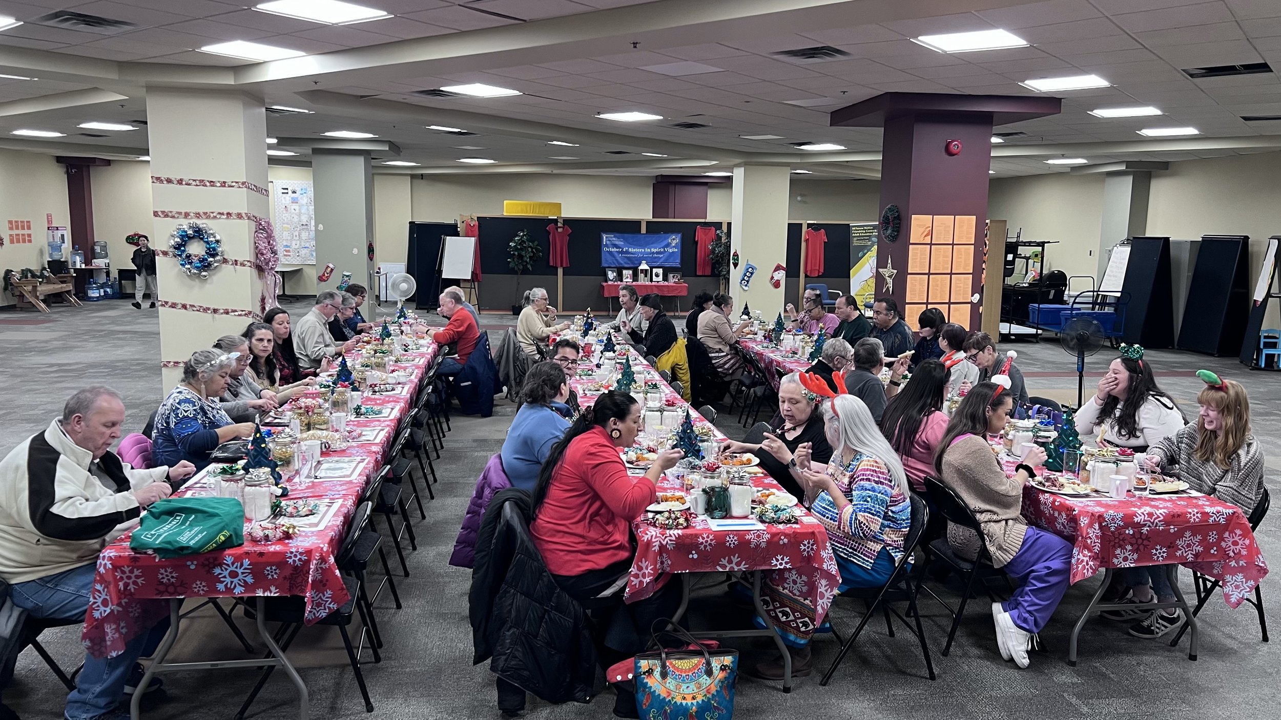 Community gathering with long tables decorated for Christmas, people wearing holiday accessories, and a stage in the background with Christmas-themed decorations.