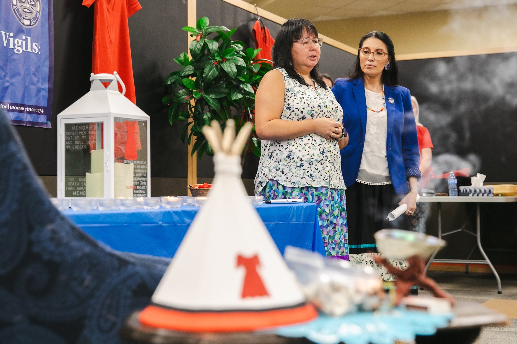 Two women speak at a gathering, with a table displaying small candles, a lantern with memorial messages, and a paper hat with a red ribbon in the foreground.