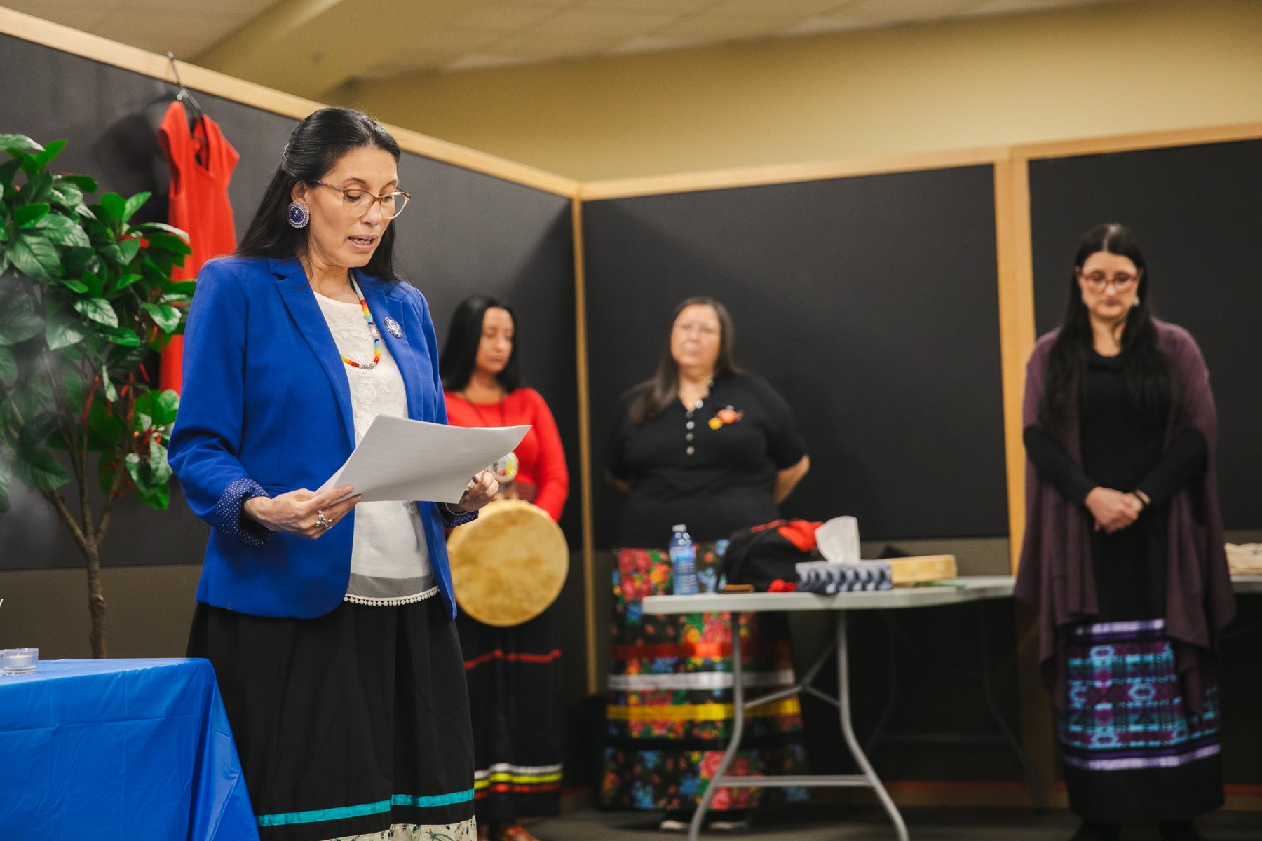 A woman in a blue blazer and glasses reads from a paper while four women in traditional Native American attire, including colorful skirts and accessories, stand behind her in a room with black walls and a table with various items.