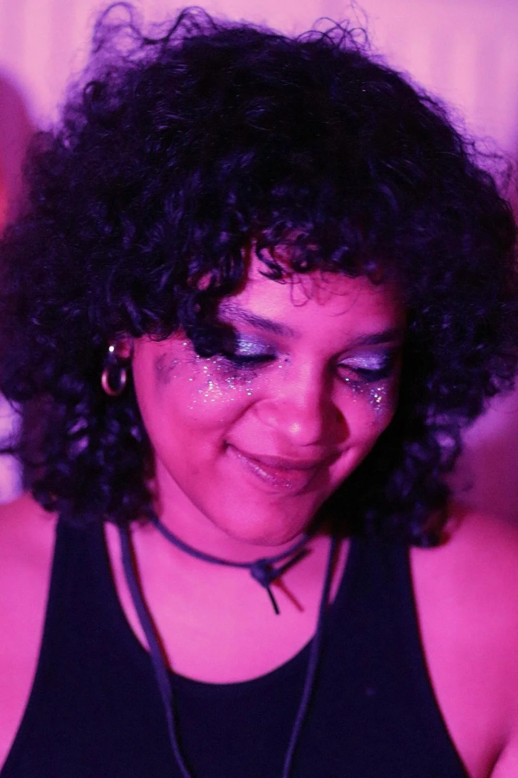 Portrait d'une femme avec des boucles noires, maquillage brillant et glitter autour des yeux, portant un choker noir et un haut sans manches noir, sous un éclairage violet