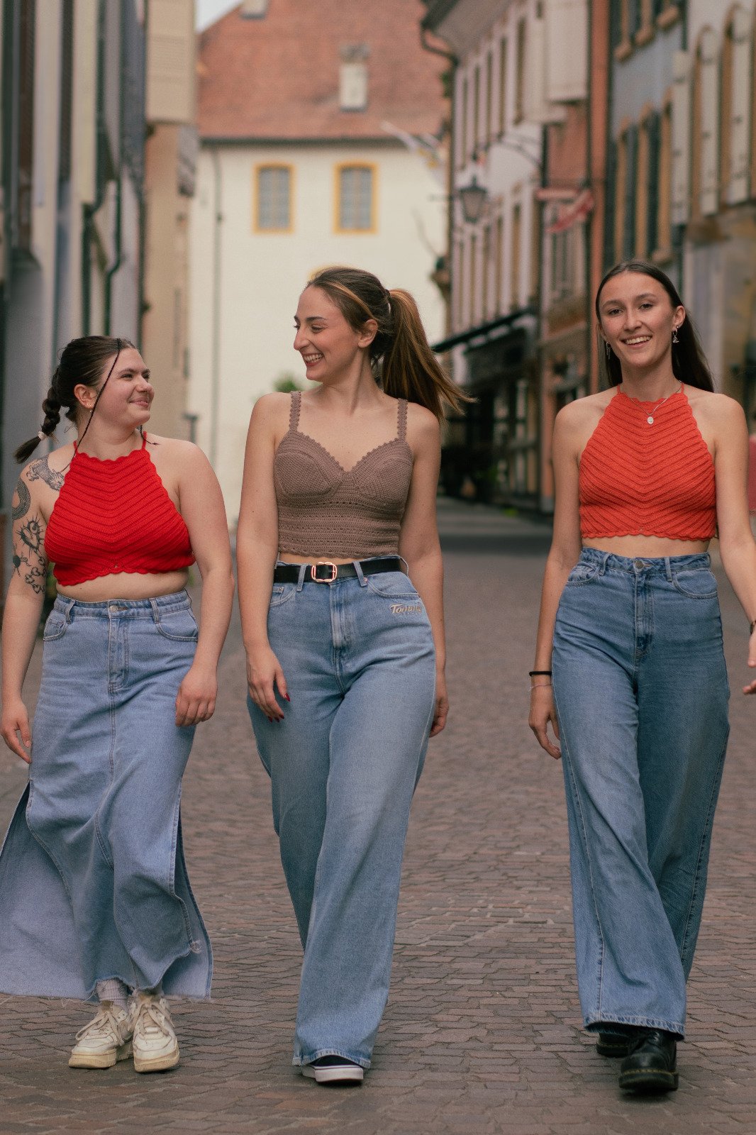 Trois jeunes femmes marchant main dans la main dans une rue pavée, souriantes et dégagées, avec en arrière-plan des bâtiments colorés typiques d'une vieille ville.