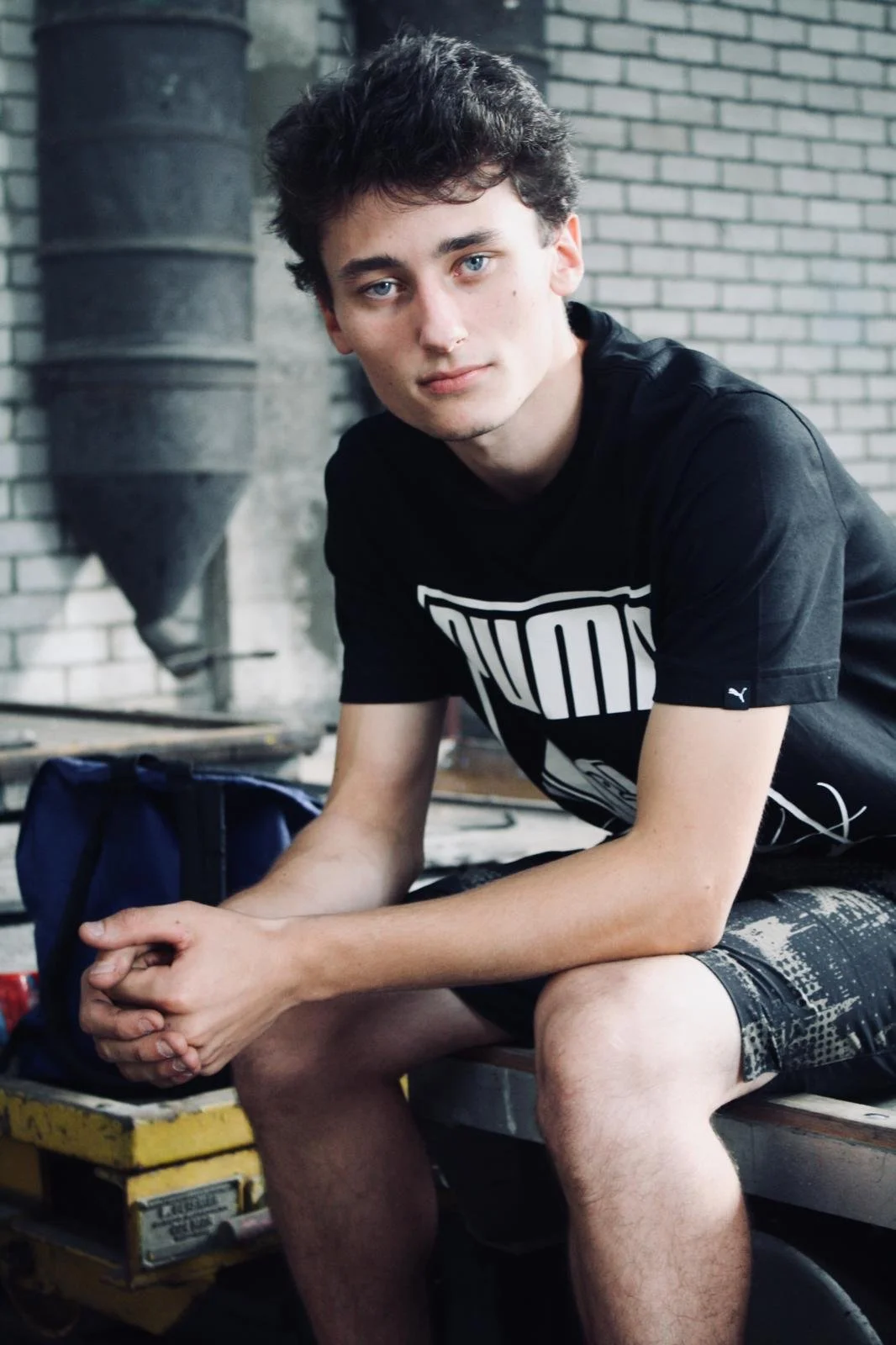 Jeune homme assis dans un environnement industriel avec un mur en briques et une cheminée en arrière-plan, portant un t-shirt noir et des shorts, regardant vers l'objectif.