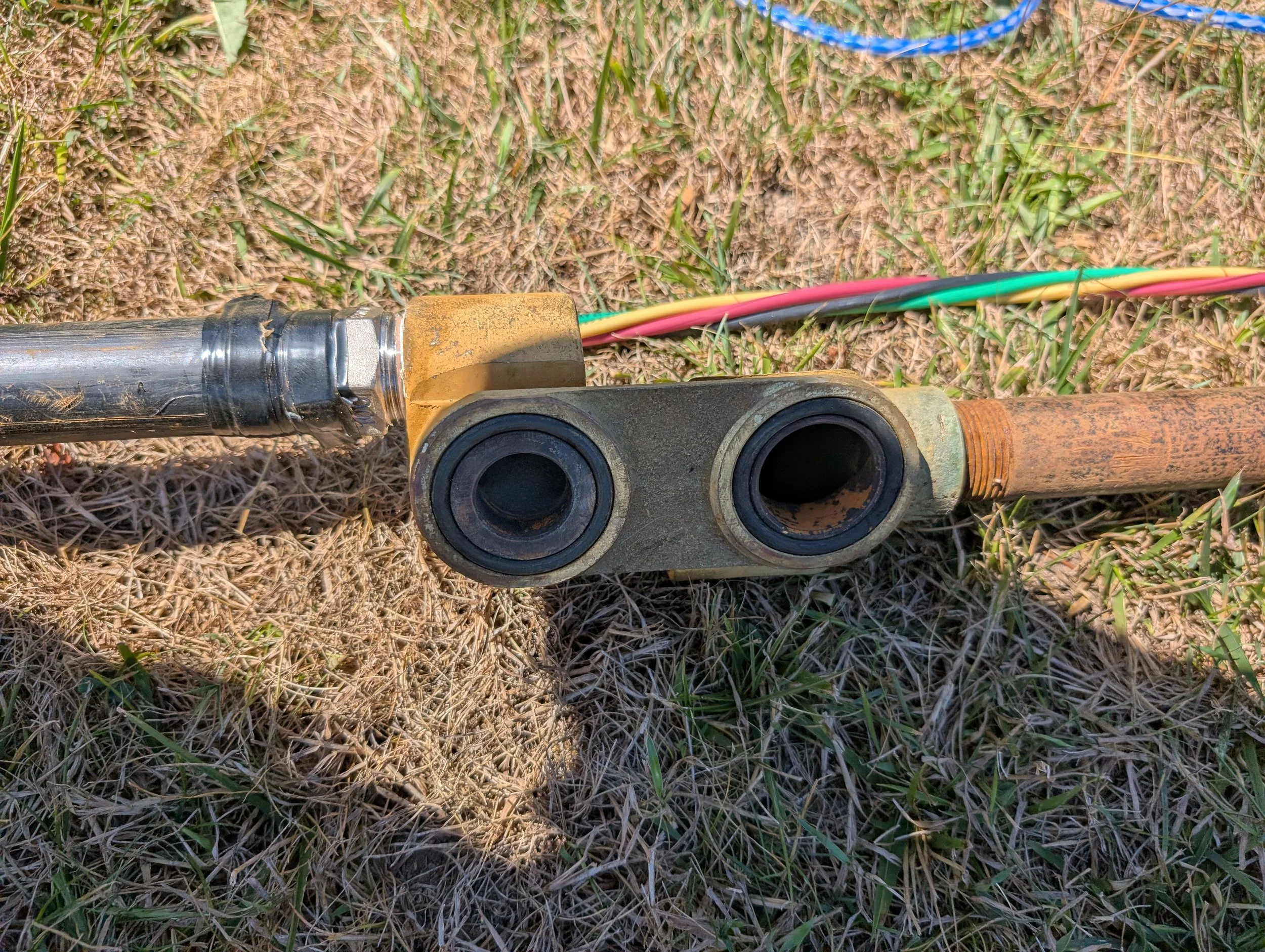 Close-up of a mechanical part on the ground, connected to multicolored wires, with grass and dirt in the background.