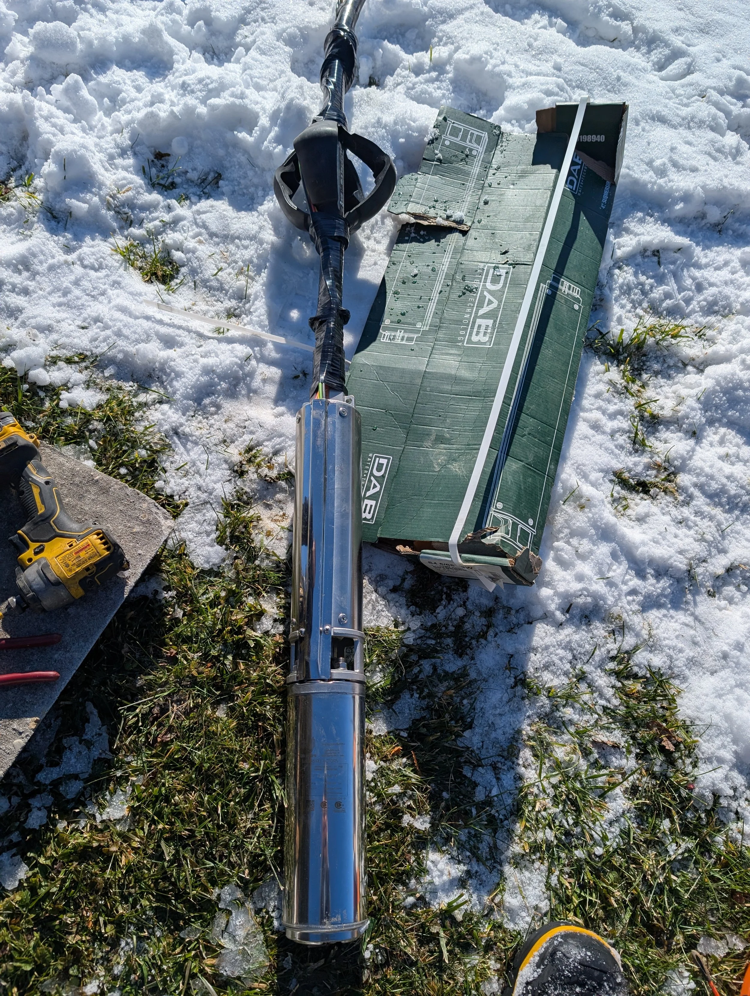 A snow-covered ground with a partially visible hand and a tool or piece of equipment, a long metal pole, a cardboard box, and a yellow and black power drill.