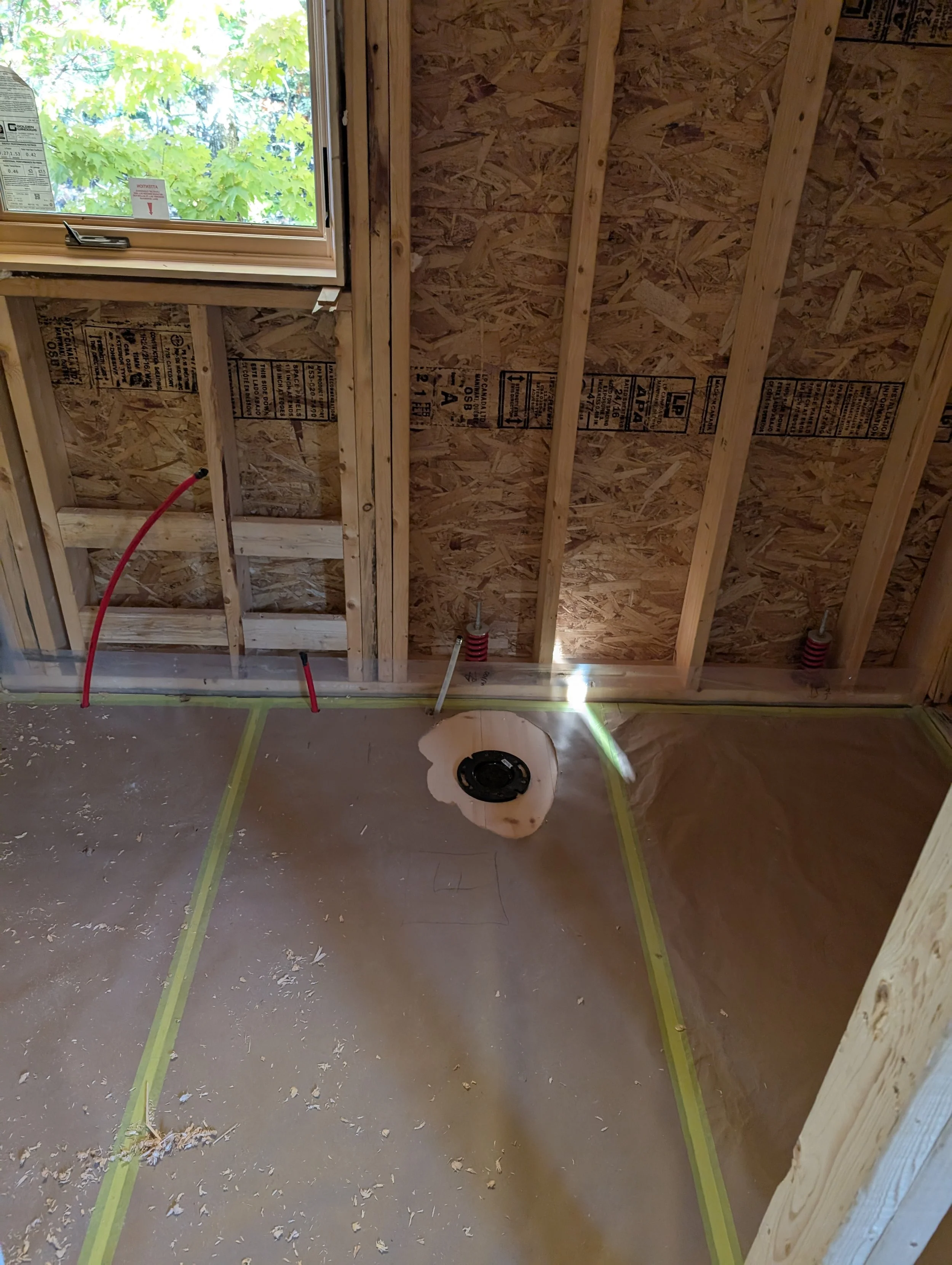 Interior of a building under construction with unfinished wood framing, a window showing green leaves outside, and a hole in the floor with a wire and a black speaker or device in the center.