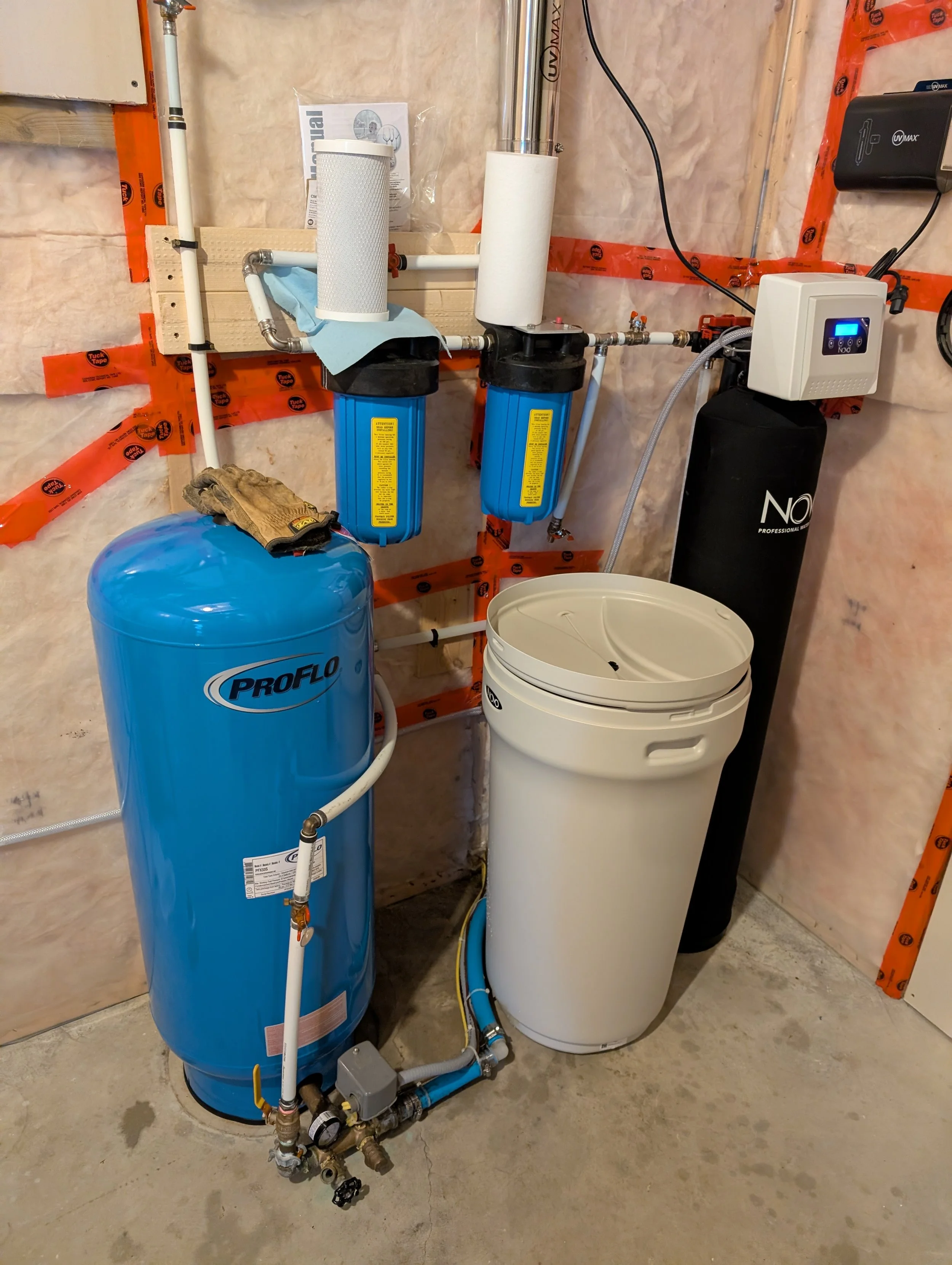 A water filtration system installed in a utility room, consisting of a large blue ProFlo water tank, a white water softener, a manual filter, and various pipes and valves.