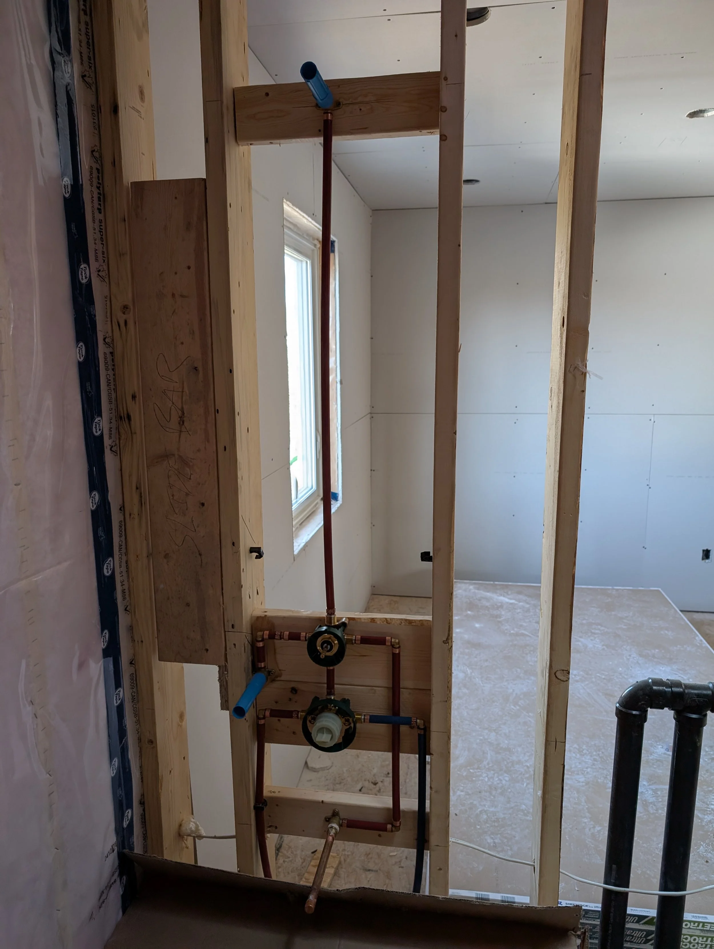 Interior view of a room under construction with exposed wooden framing and plumbing pipes. There is a window on the wall and no ceiling finished yet.