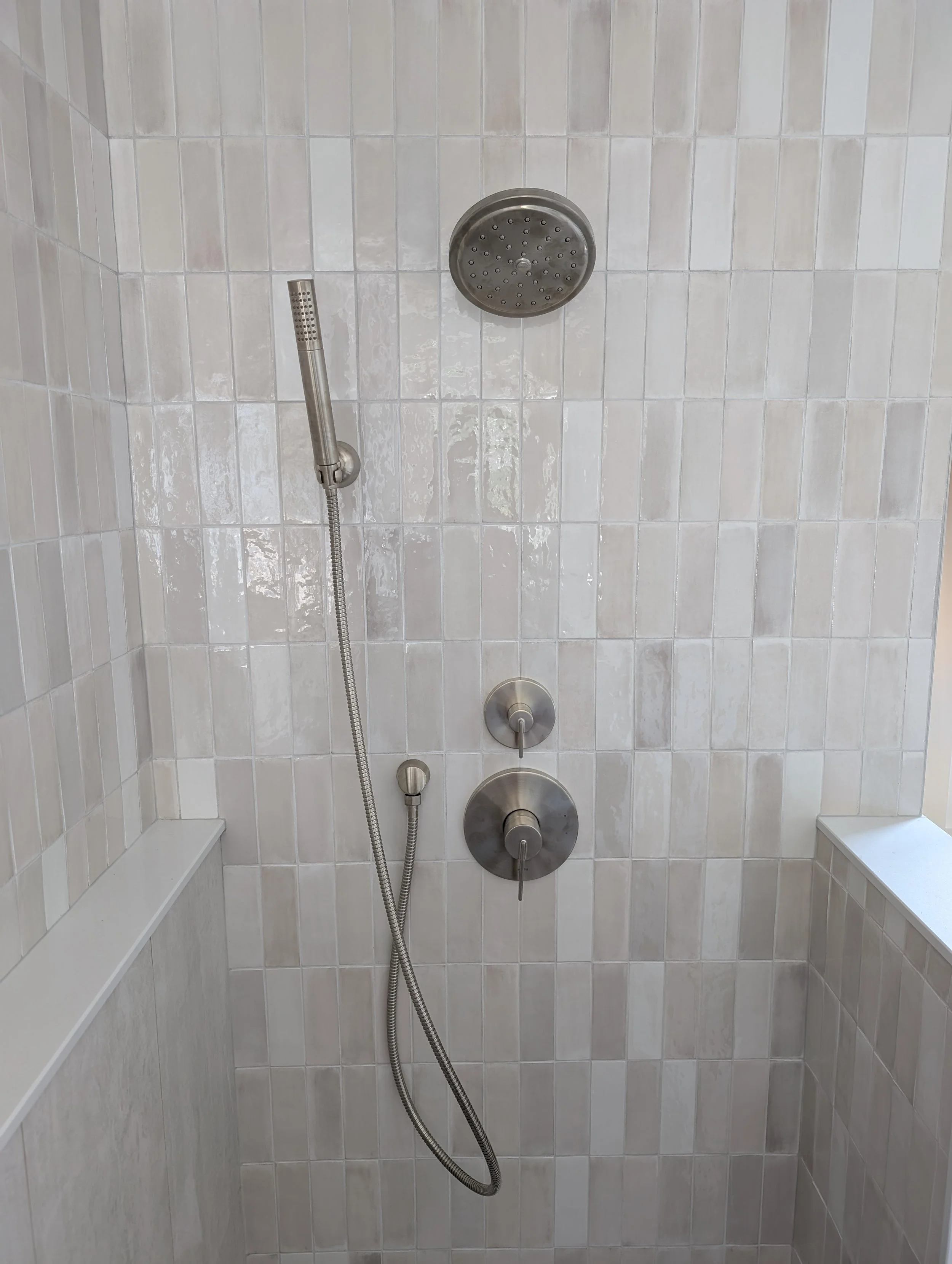A walk-in shower with beige tiled walls, a black showerhead, a handheld shower wand, and two control knobs.