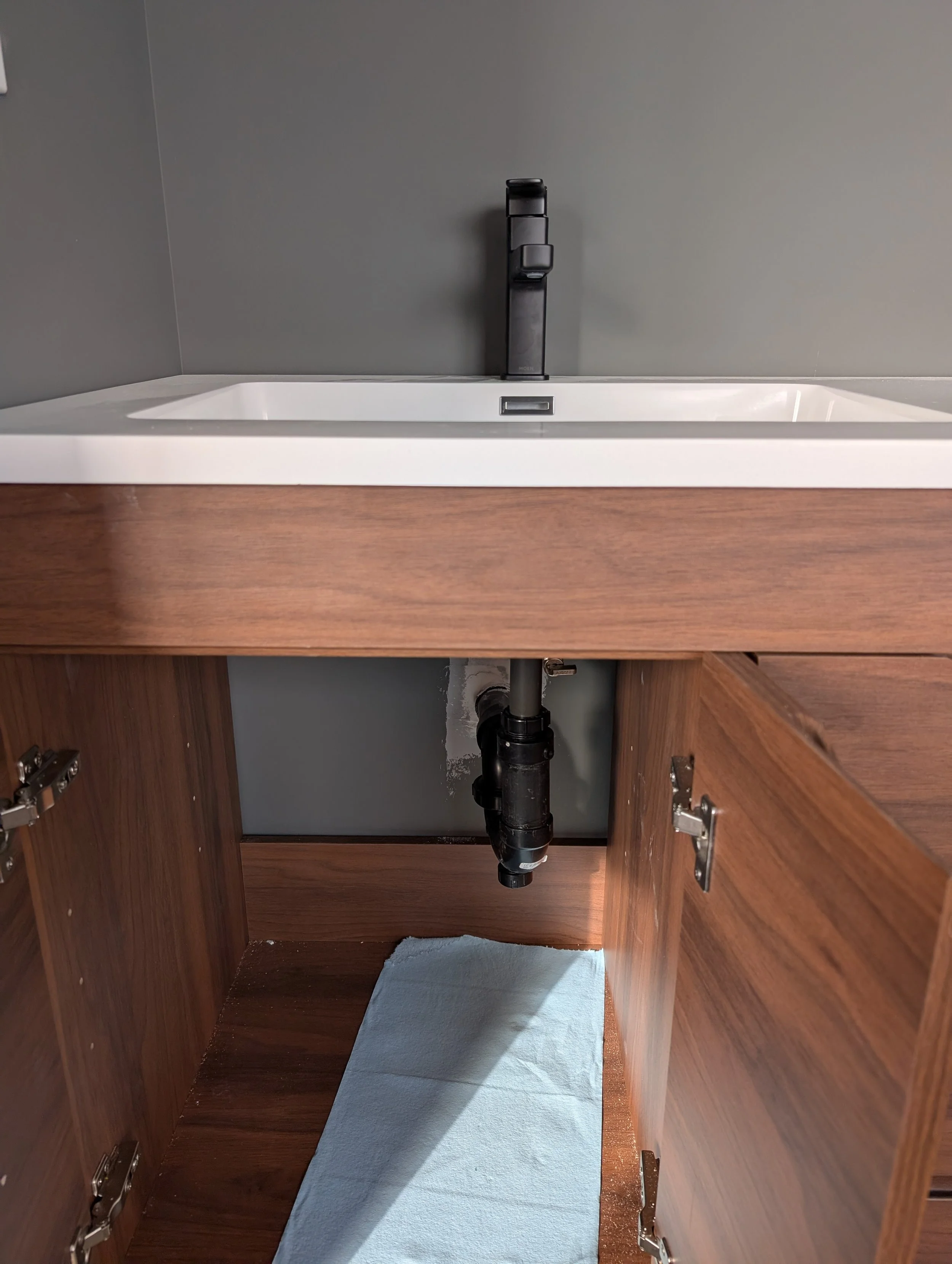 A kitchen cabinet open beneath a sink, showing plumbing pipes, with a blue mat on the floor and a gray wall behind.
