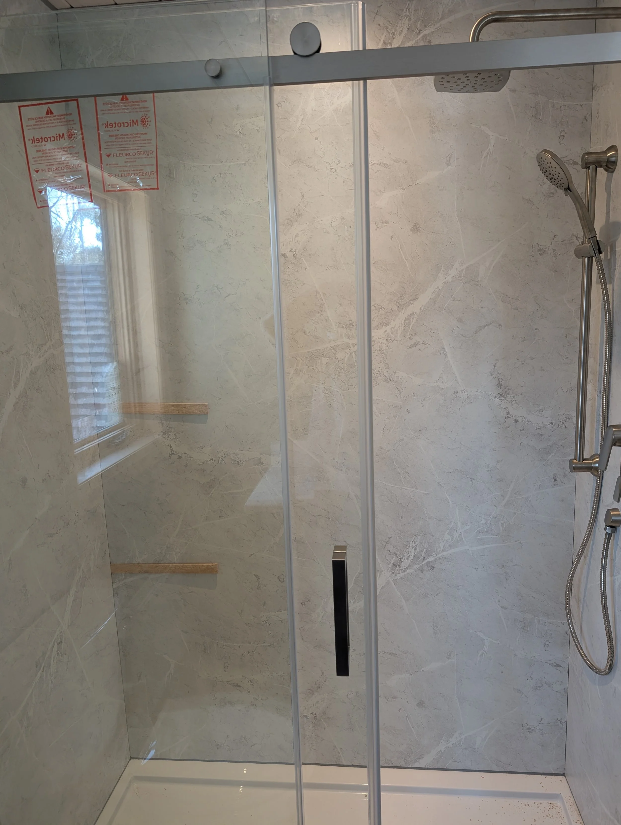 Shower with a glass door, beige marble walls, a round showerhead attached to a vertical bar, and a handheld showerhead on a flexible hose. There are two red and white warning labels on the inside of the glass door, and a reflection of a window with b