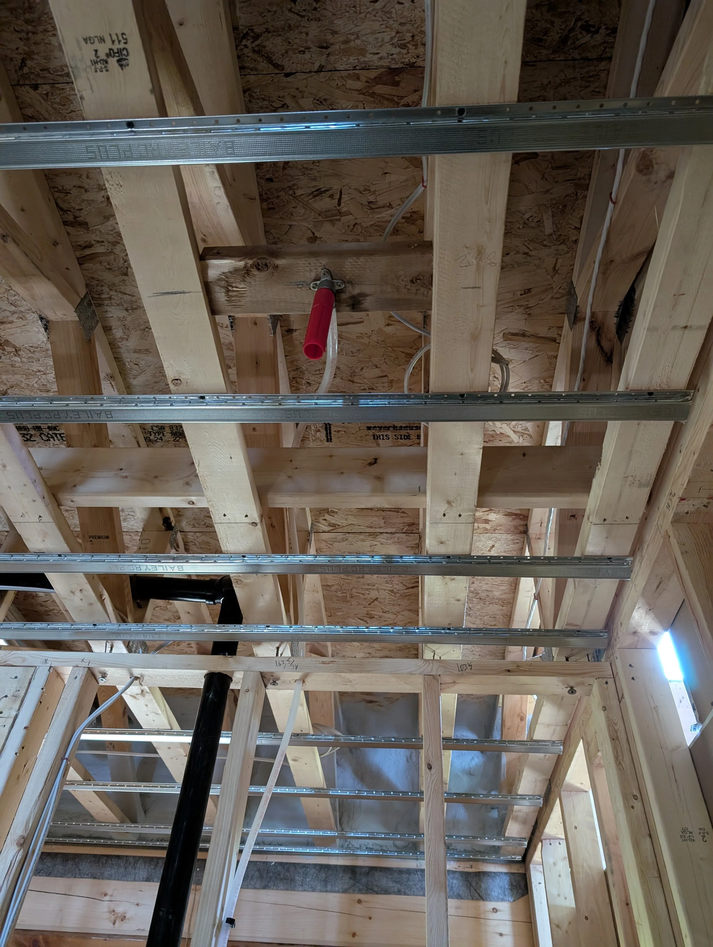 Interior view of a building under construction with exposed wooden framing, metal support beams, electrical wiring, and plumbing pipes.