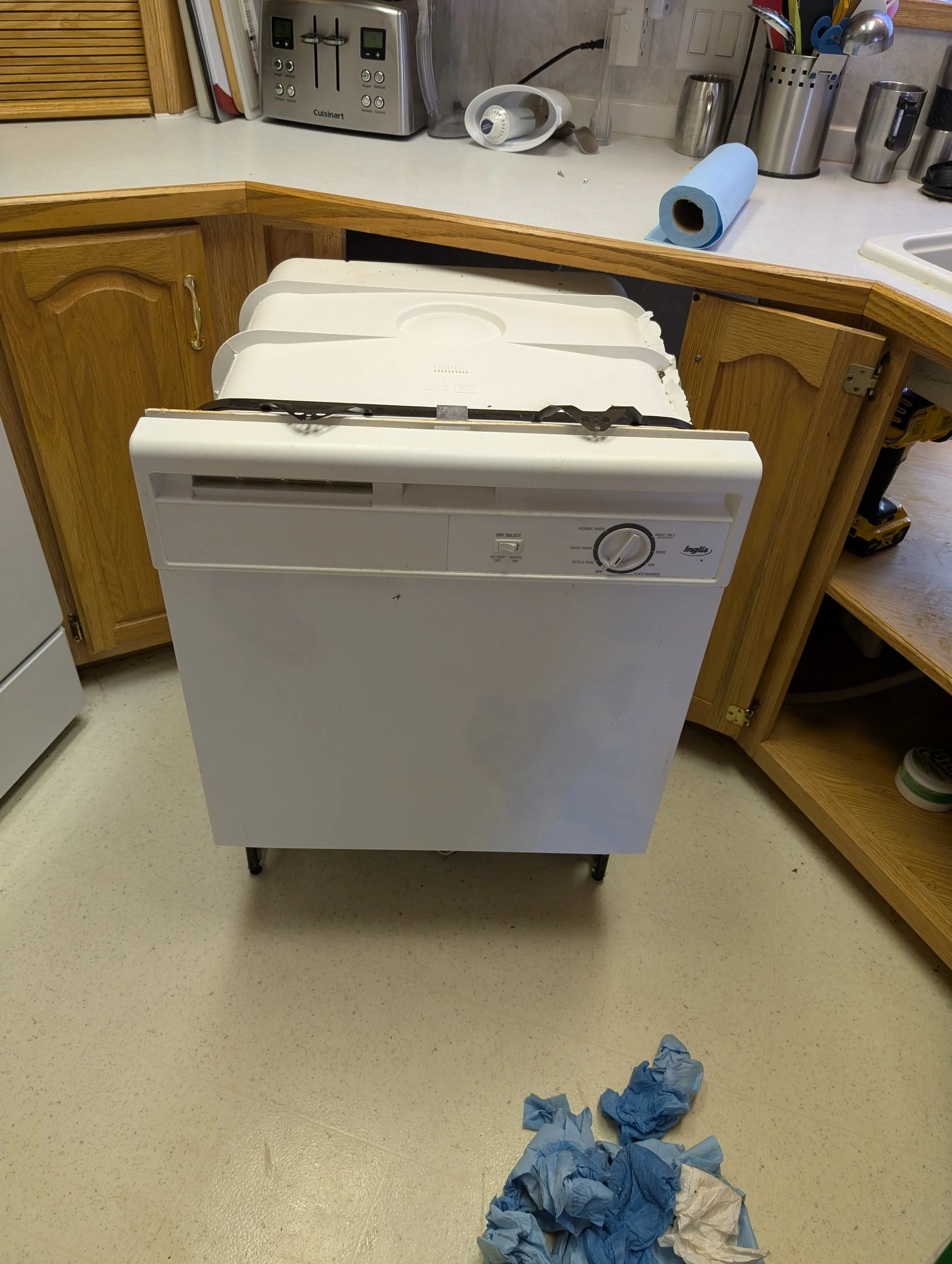 A dishwasher with its front panel removed and crumpled blue and white paper towels on the floor in front. The kitchen has a wooden cabinet and various small appliances on the counter, such as a toaster, a paper towel roll, and utensils.