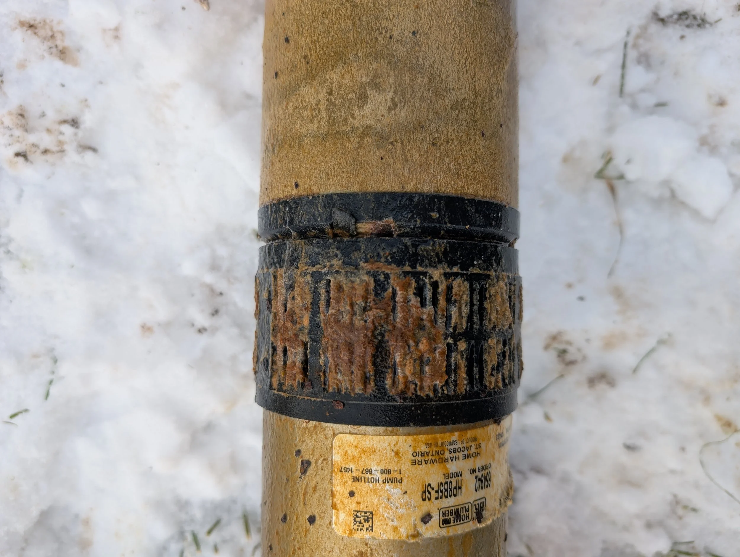 Close-up of a dirty, rusted pipe with a yellow label partially visible, resting on snow-covered ground.