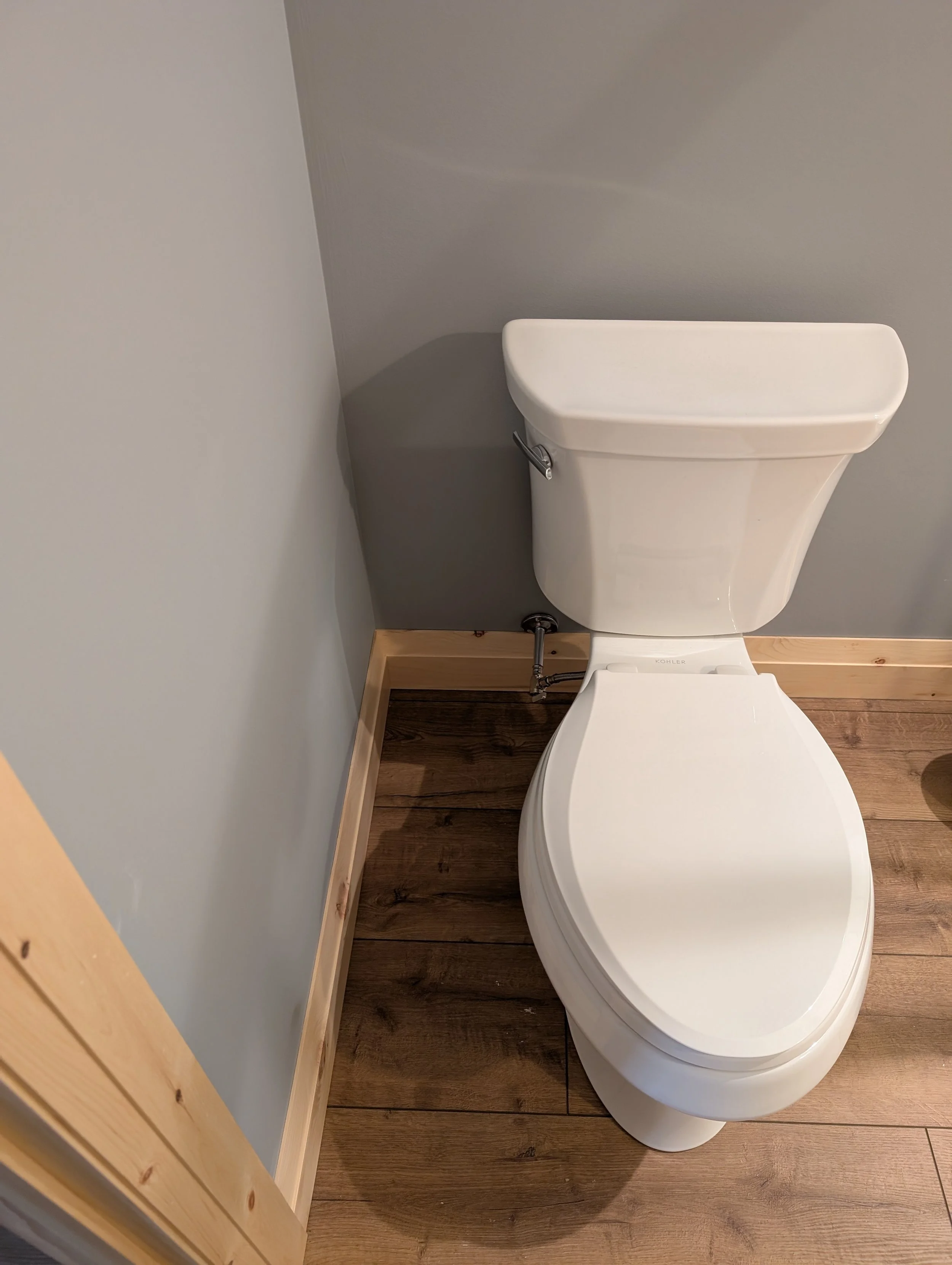 A modern white toilet with a tank and a flush handle, installed next to a corner with light gray walls and wooden flooring.