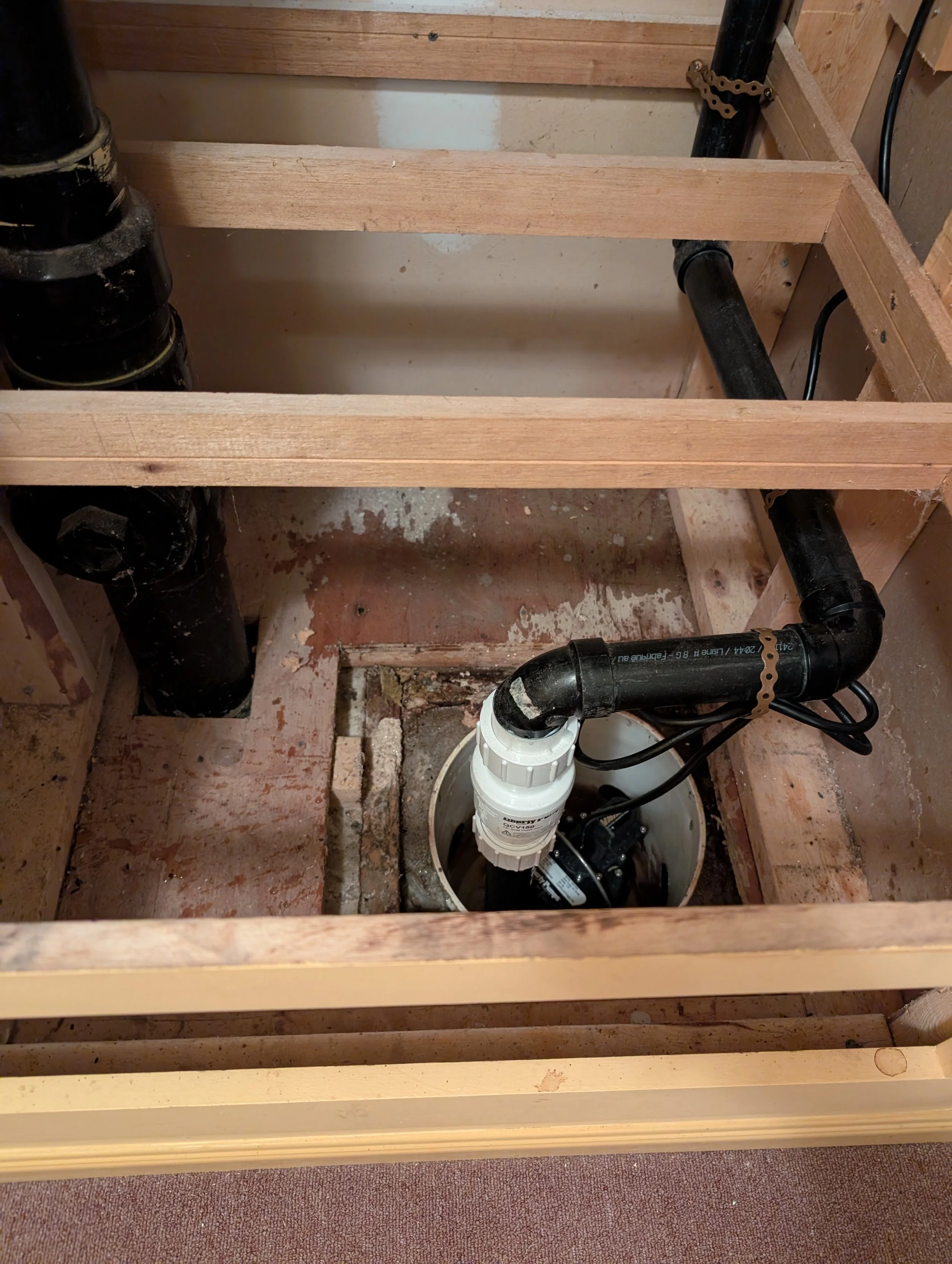 View of a bathroom wall with plumbing pipes, including black drain pipes and white PVC pipes, installed through an open wooden framing structure. The environment appears to be under construction or renovation.