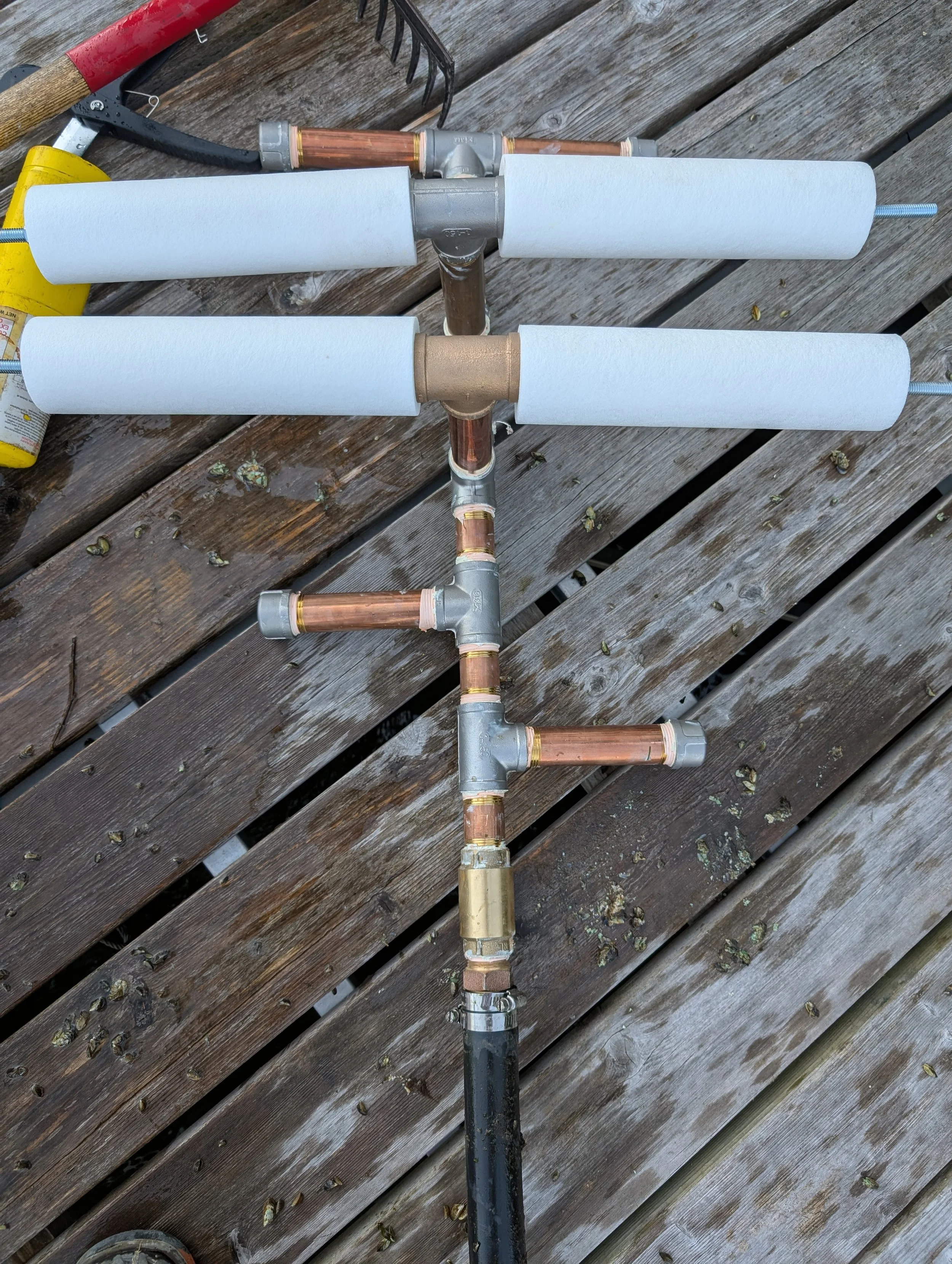 A makeshift DIY sprinkler system made from PVC pipes and copper fittings laid on a wooden deck.