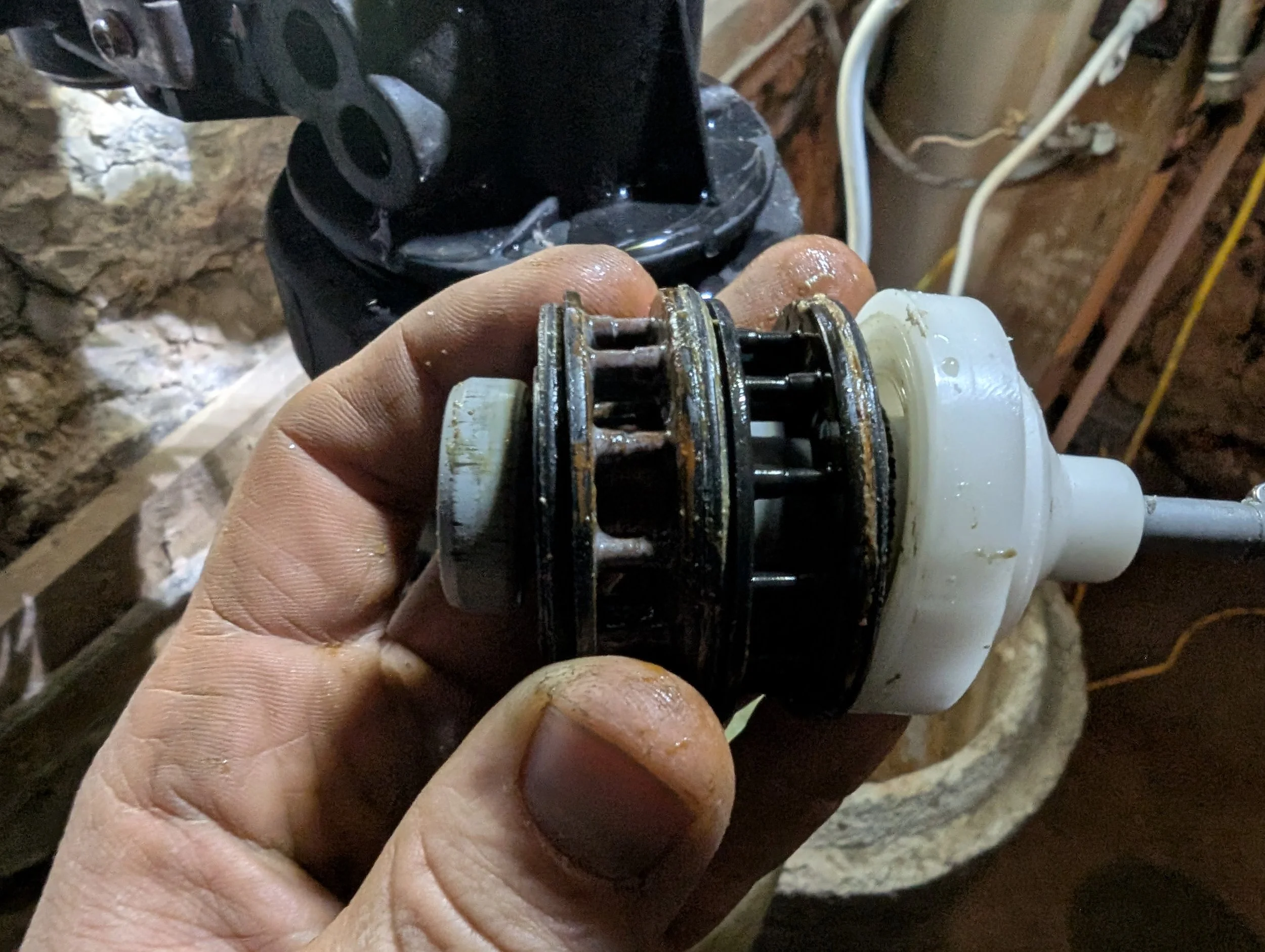 A person's hand holding a small mechanical part with rust, dirt, and grease, in front of other equipment in a workshop or garage.