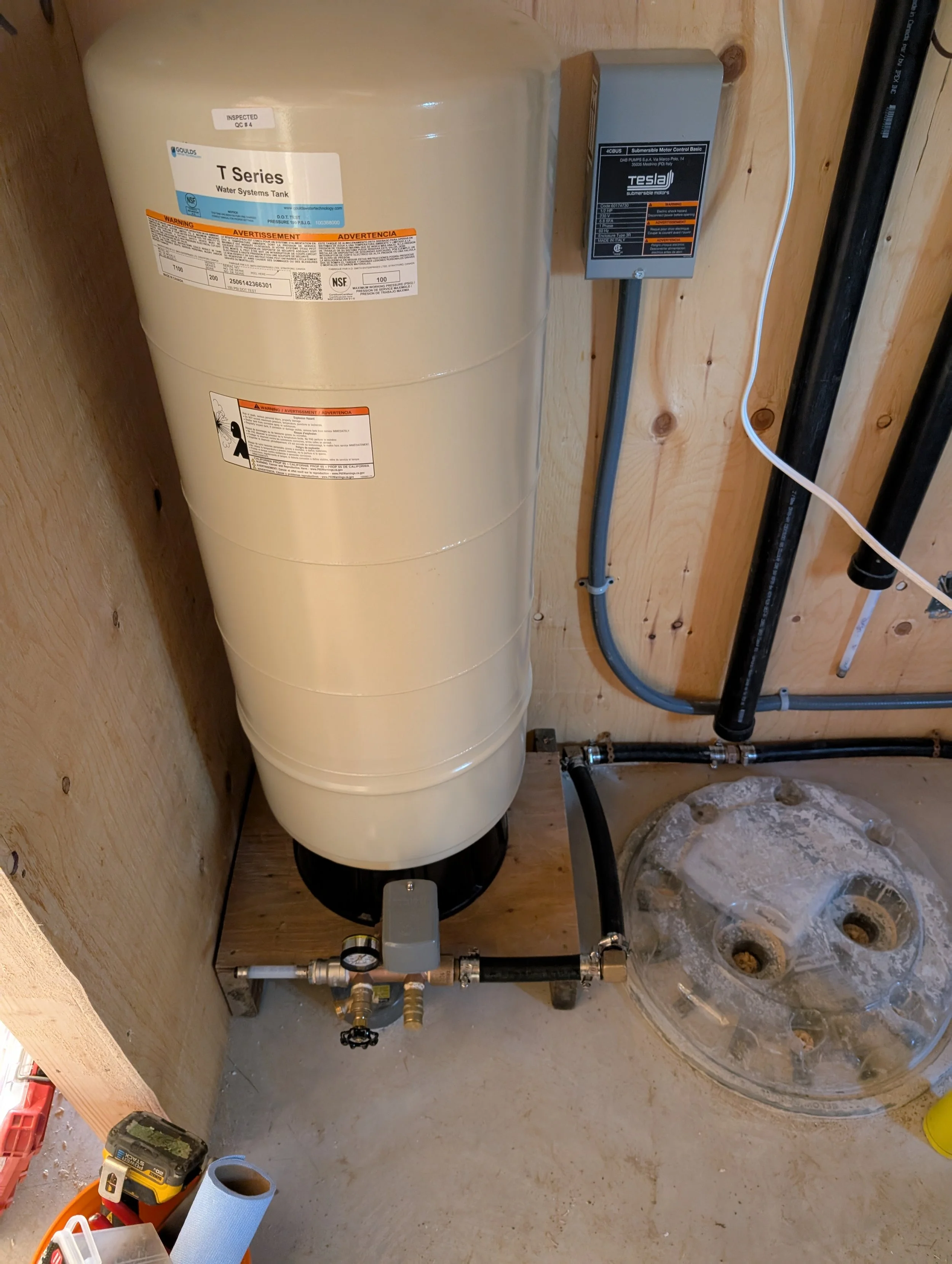 Installment of a water system with a large water tank, plumbing pipes, and a control box in a wooden room under construction.