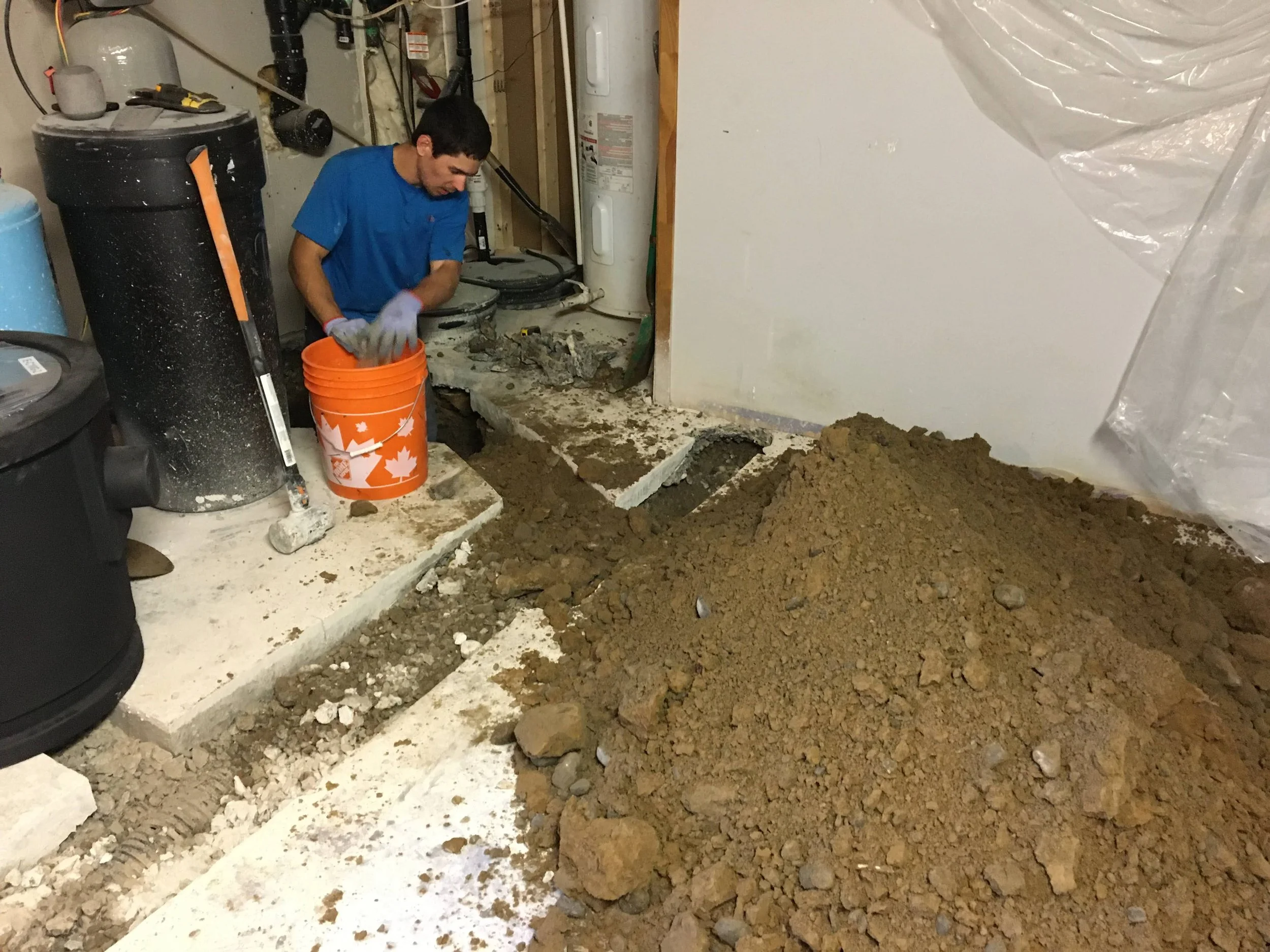 A plumber engaged in home repair, working with soil and concrete in a basement or utility room, with tools and equipment around.