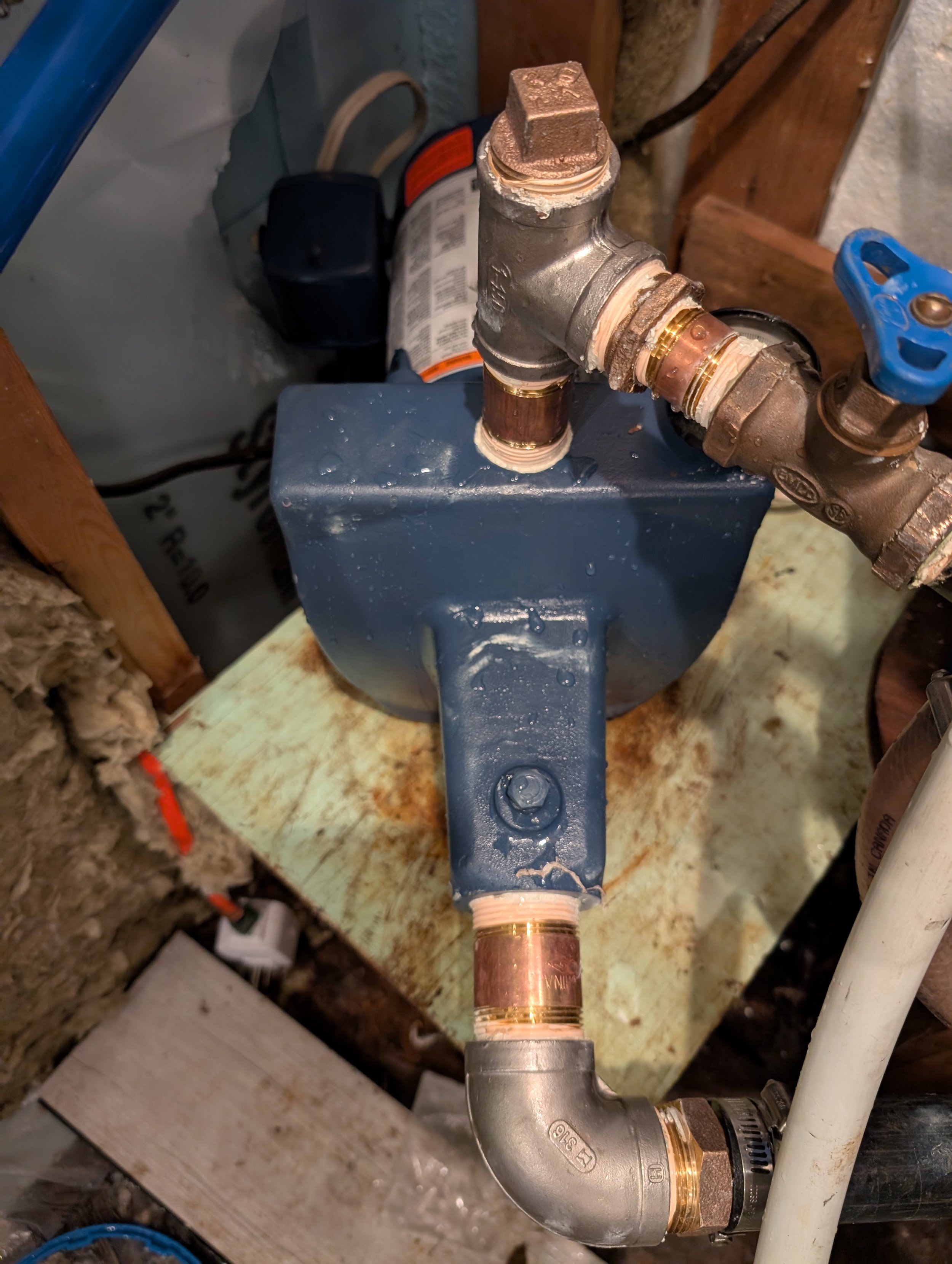 Piping system with copper and brass fittings, including a valve with a blue handle, connected to a blue box, in a construction or plumbing area with insulation and wood framing in the background.