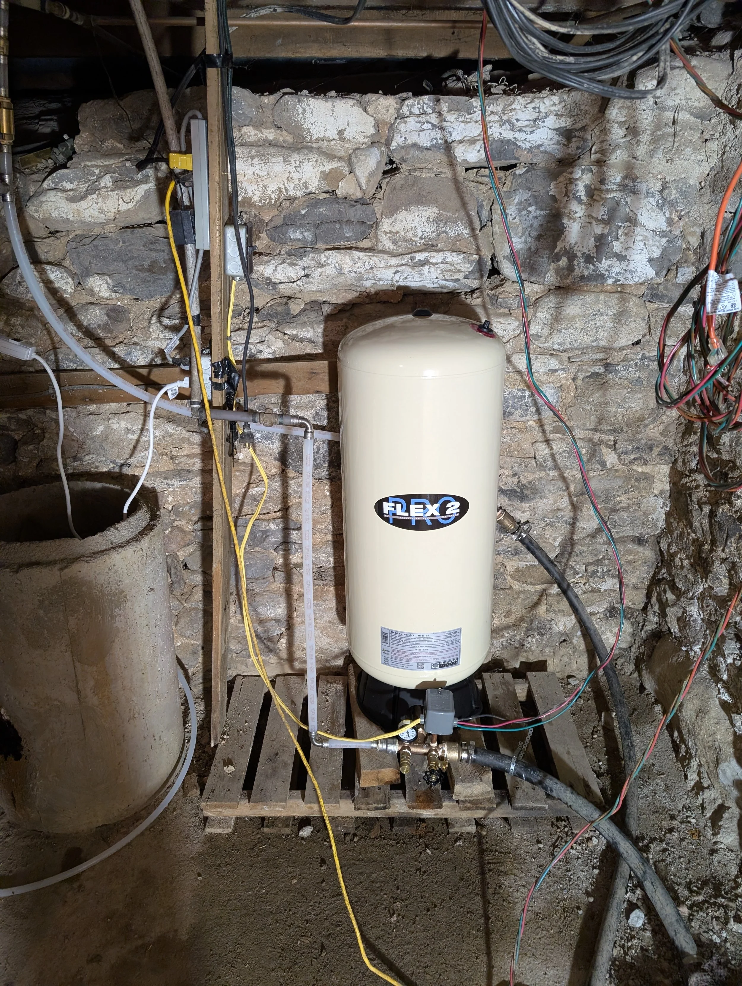 An indoor utility space with a stone wall and a white Flexcon water heater tank, various pipes, wires, and electrical components are connected around the tank, which is placed on a wooden pallet.