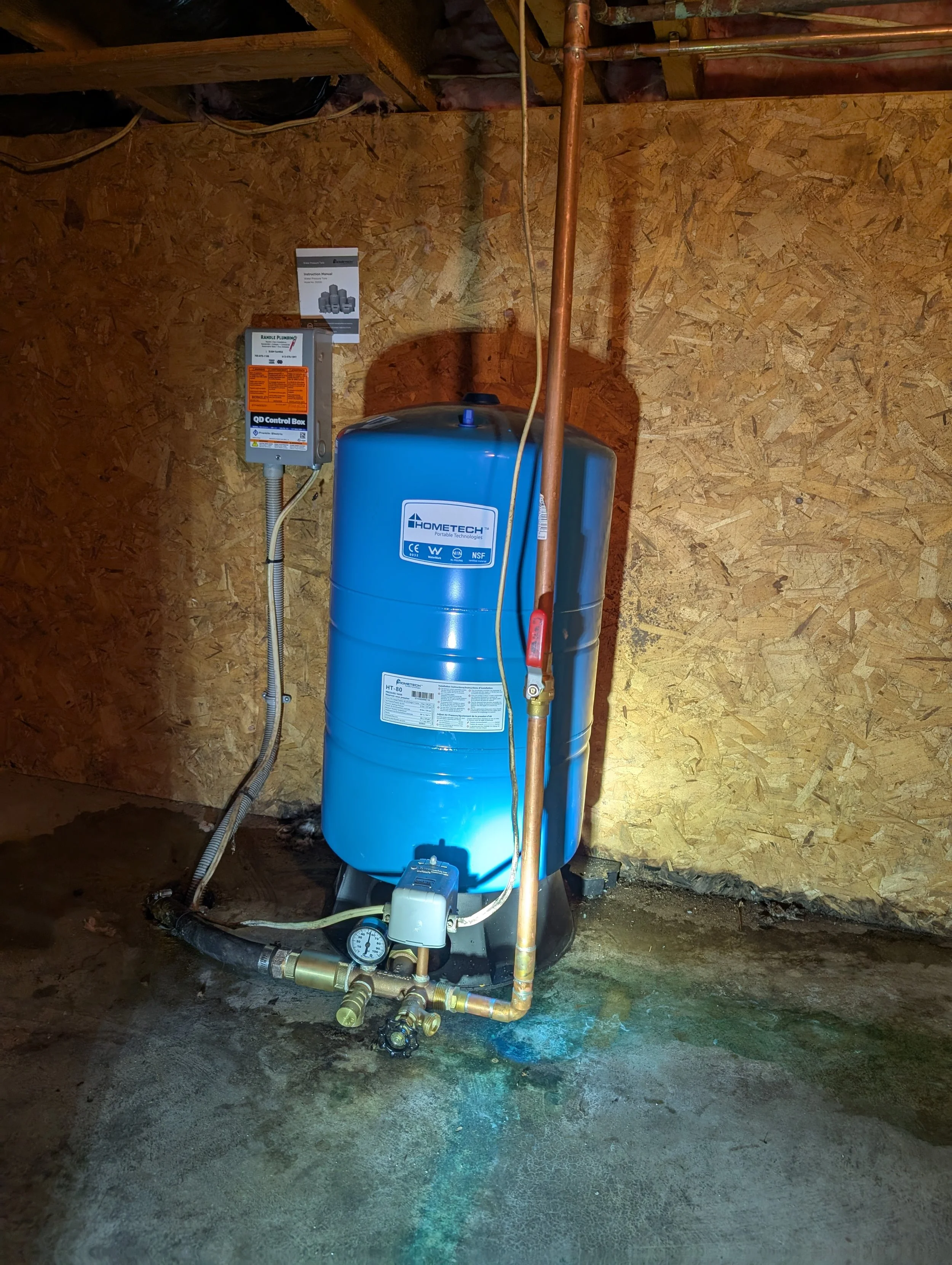 A water pressure tank with plumbing and control components in a room with plywood walls and concrete floor.