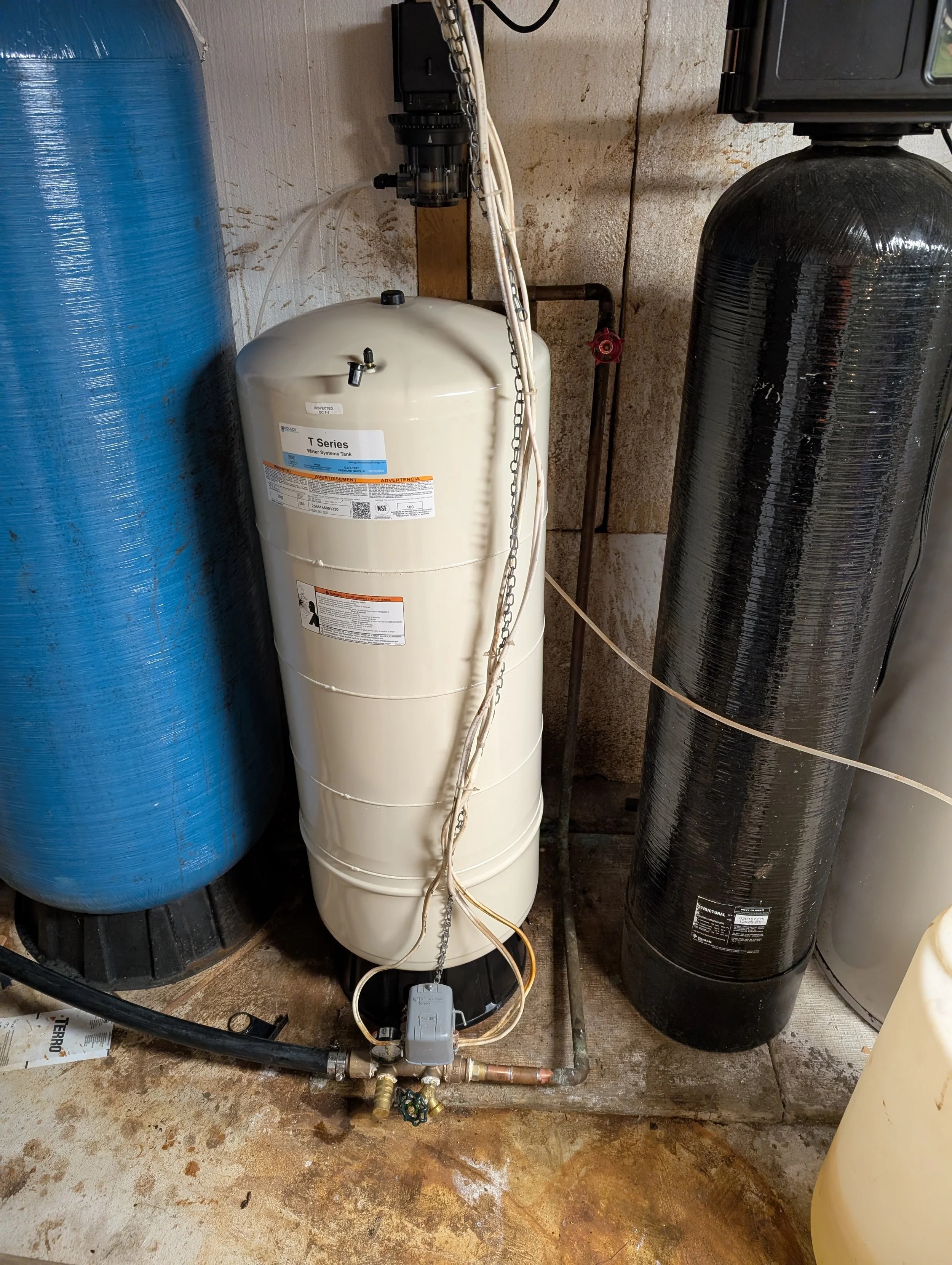 A water filtration system with a blue tank on the left, a white tank in the middle labeled 'T Series Water Systems Tank,' and a black tank on the right. Various pipes, valves, and wiring connect the tanks, installed against a concrete wall with a rus