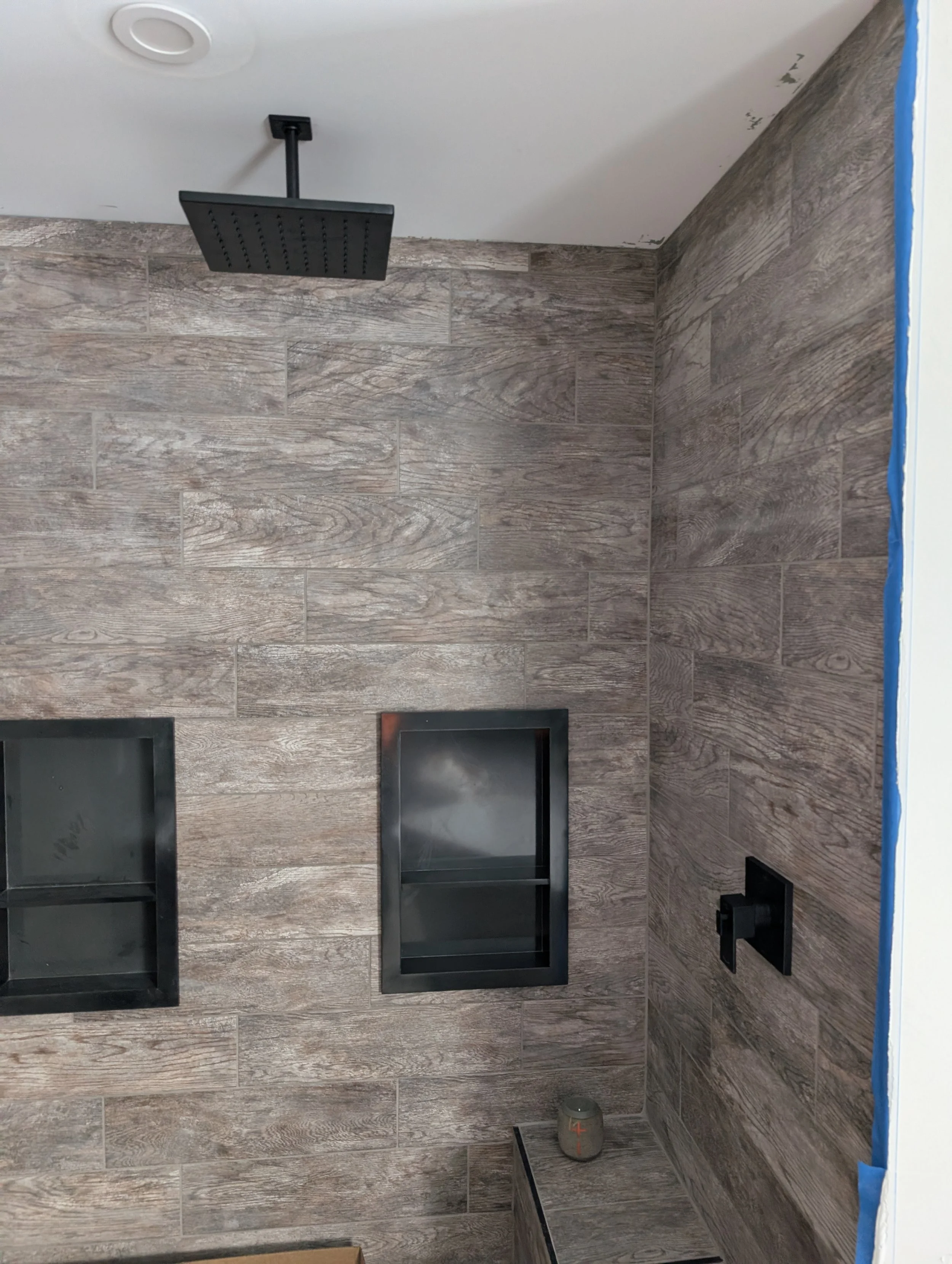 A bathroom shower area with wooden tile walls, a black square showerhead mounted on the ceiling, black framed glass shower doors, and a small gray speaker with orange crosses on a black ledge.