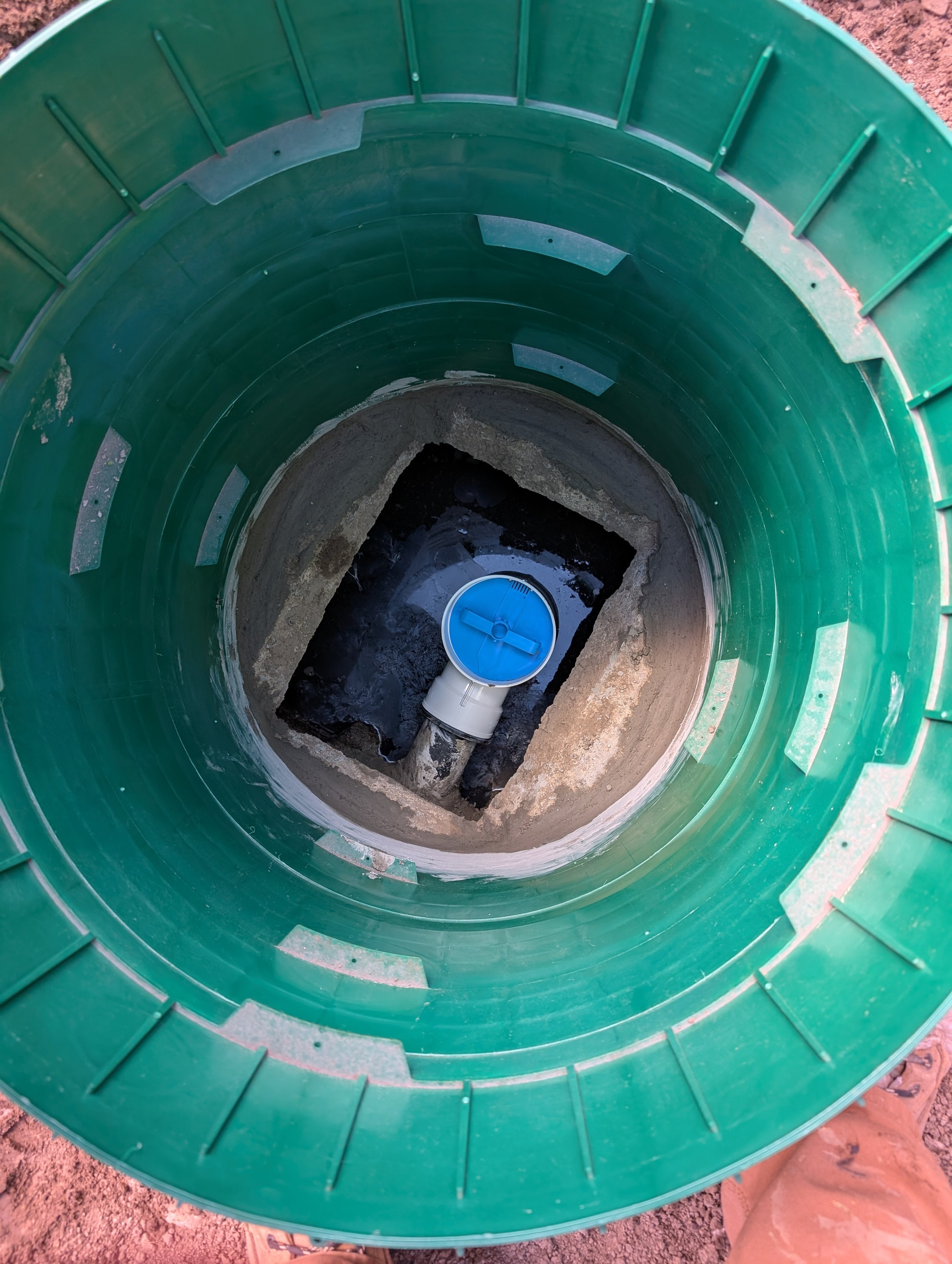 A green cylindrical septic tank or manhole with an open top revealing a blue cover and some water or sludge at the bottom, set in a dirt area.