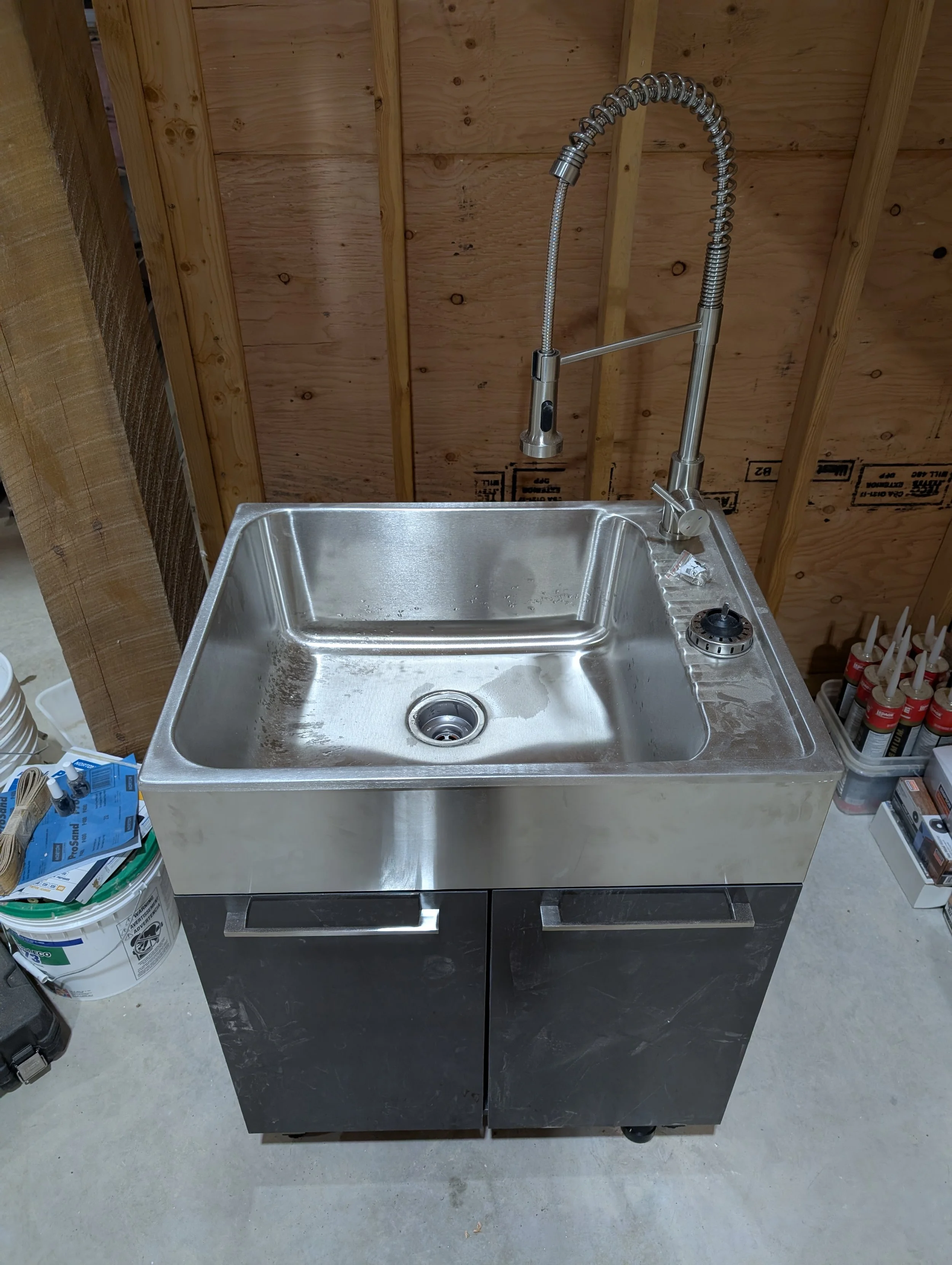 Stainless steel kitchen sink with a high-arc pull-down faucet installed, against a wooden wall with visible framing in a construction area.