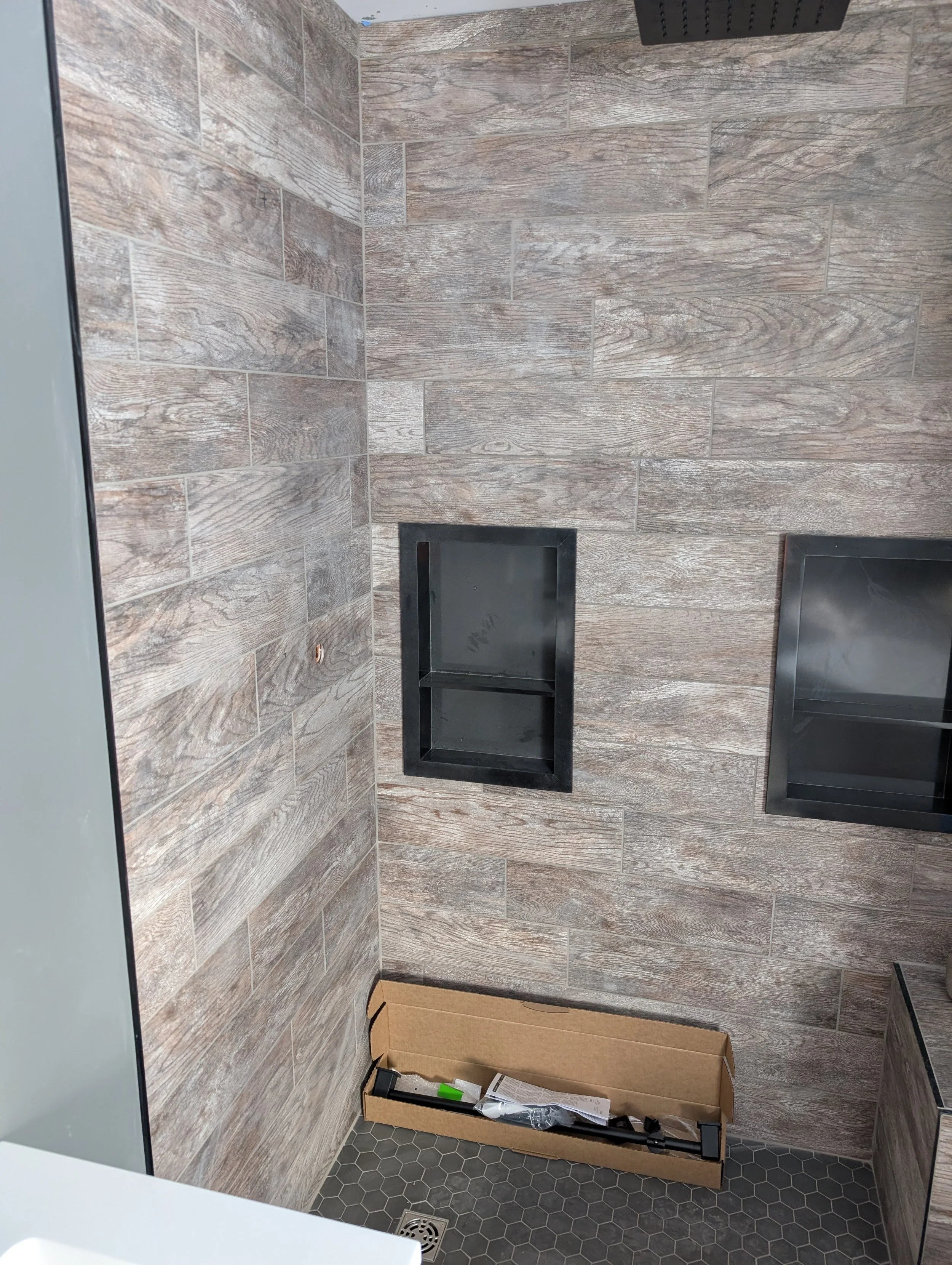 A newly constructed shower area with wood-look tile walls and black framed glass shelves. There is a black showerhead at the top. At the bottom, a cardboard box contains tools and hardware, and a black floor drain is visible.