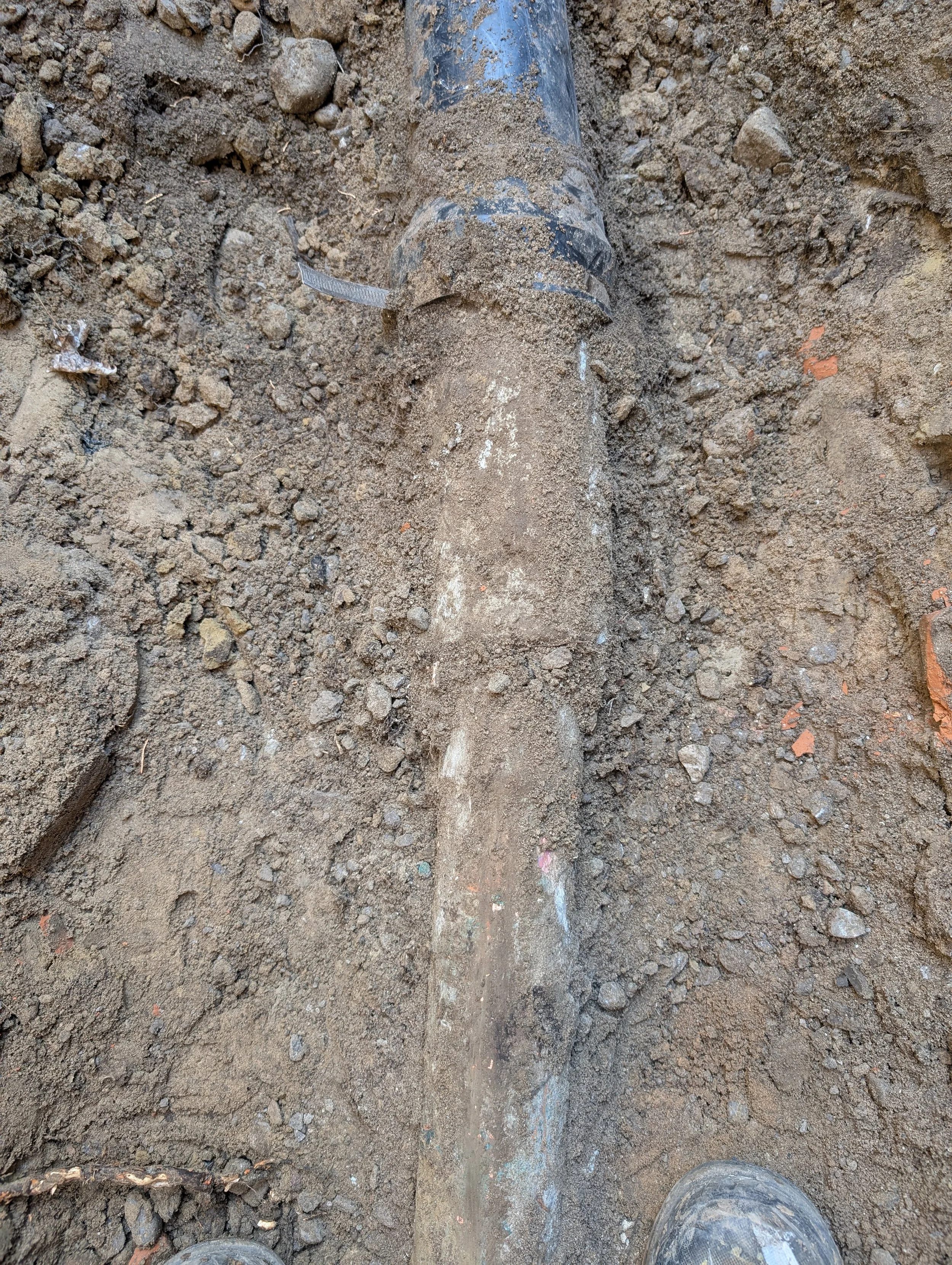 Close-up of dirt ground with a partially buried black pipe and some measuring tape, with a person's muddy boots visible at the bottom.