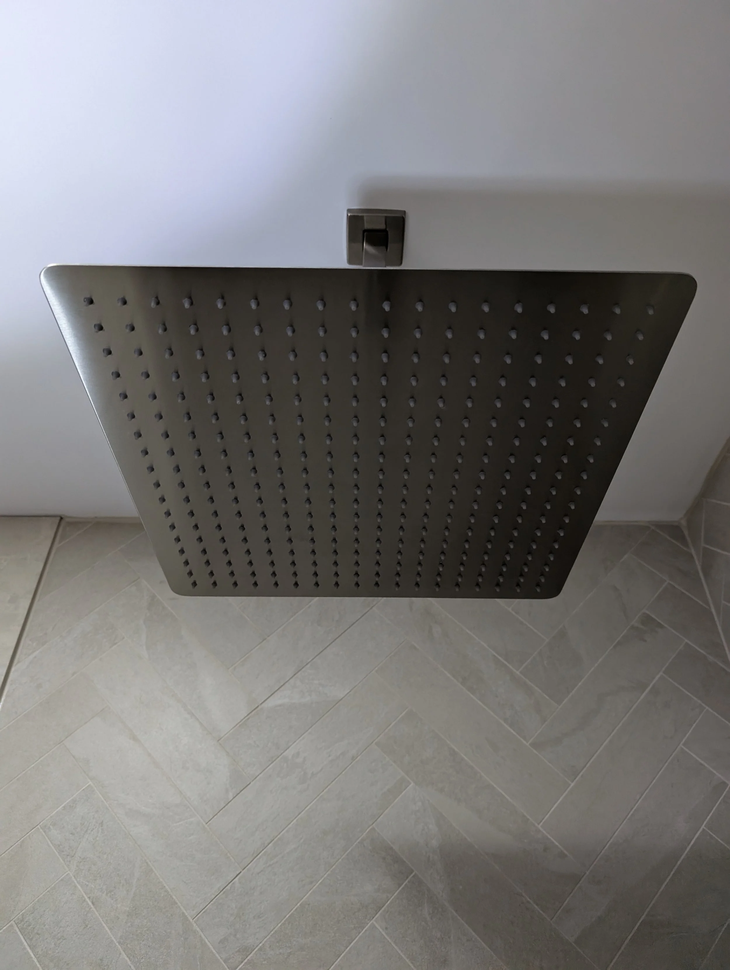 A modern rainfall showerhead with a square shape mounted on a white ceiling, with a gray square control on the ceiling, and beige tile flooring below.